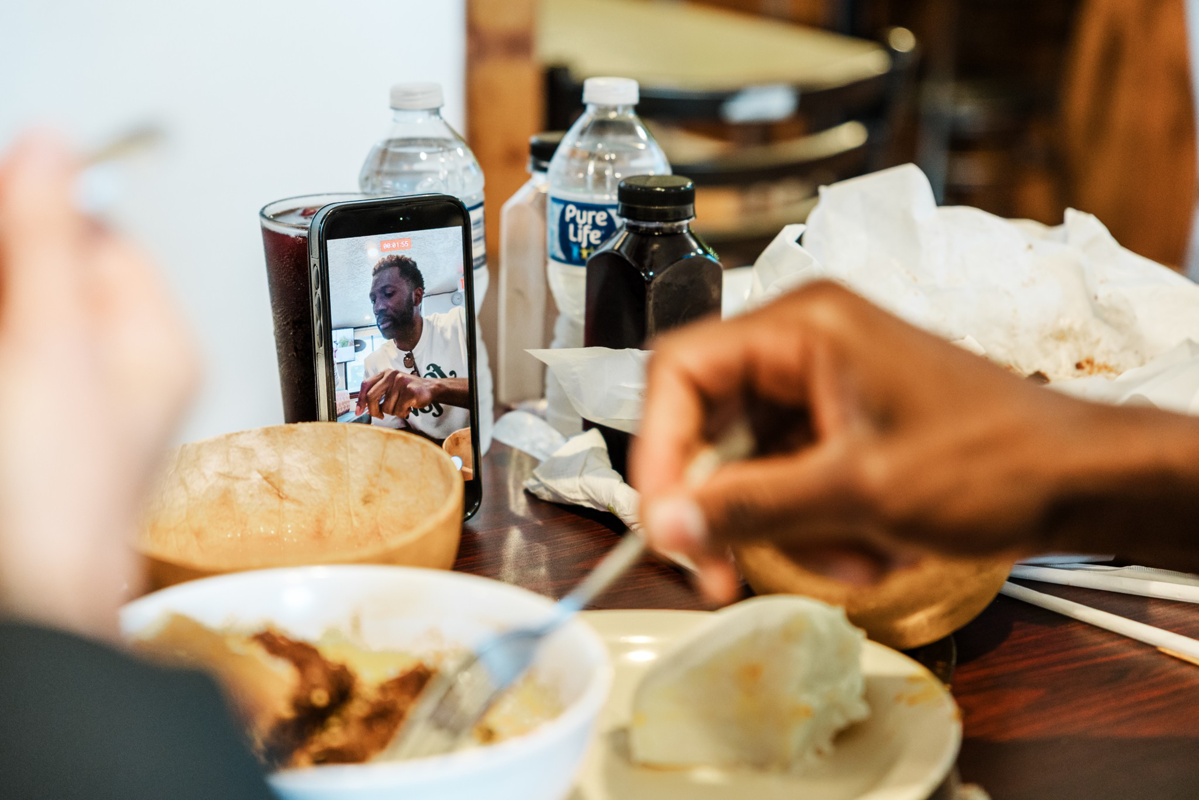A phone on a table displays a video call with a man, while two people eat a meal including bread and stew, with water bottles and containers nearby.