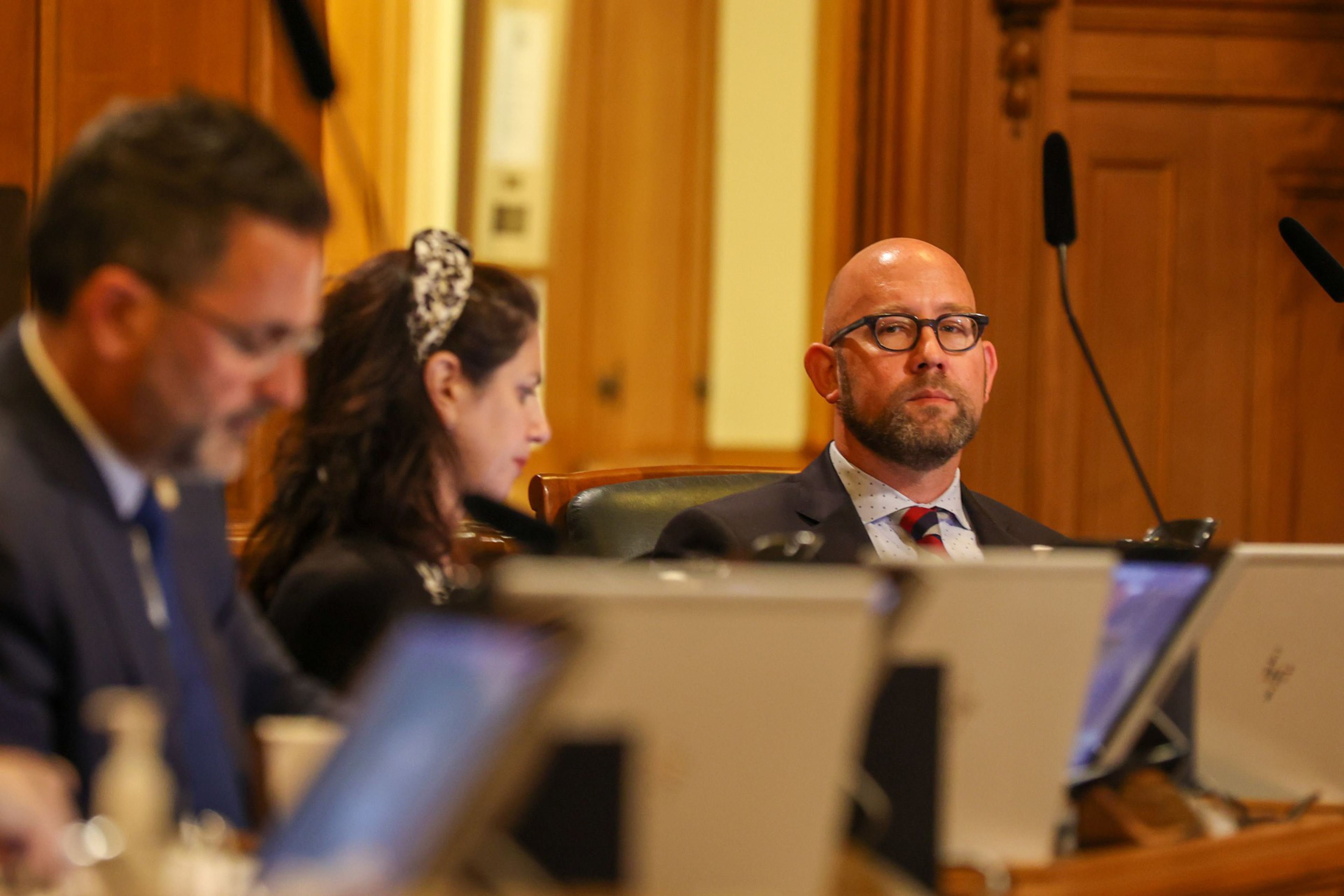 Three people are seated in a formal setting with laptops in front of them; one bald man with glasses looks directly ahead while the others focus on their screens.
