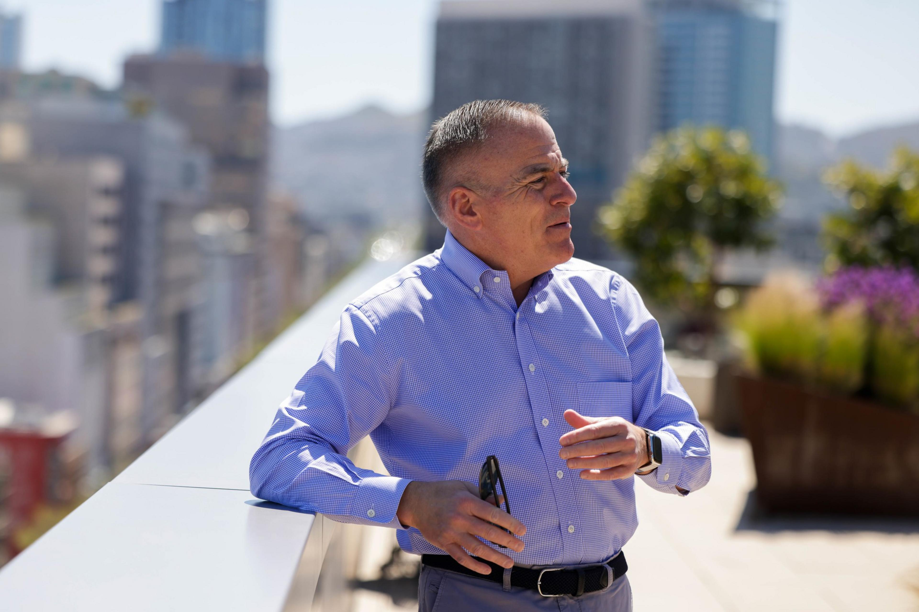 A man in a blue shirt stands outside with a cityscape behind him, holding sunglasses and a smartphone.