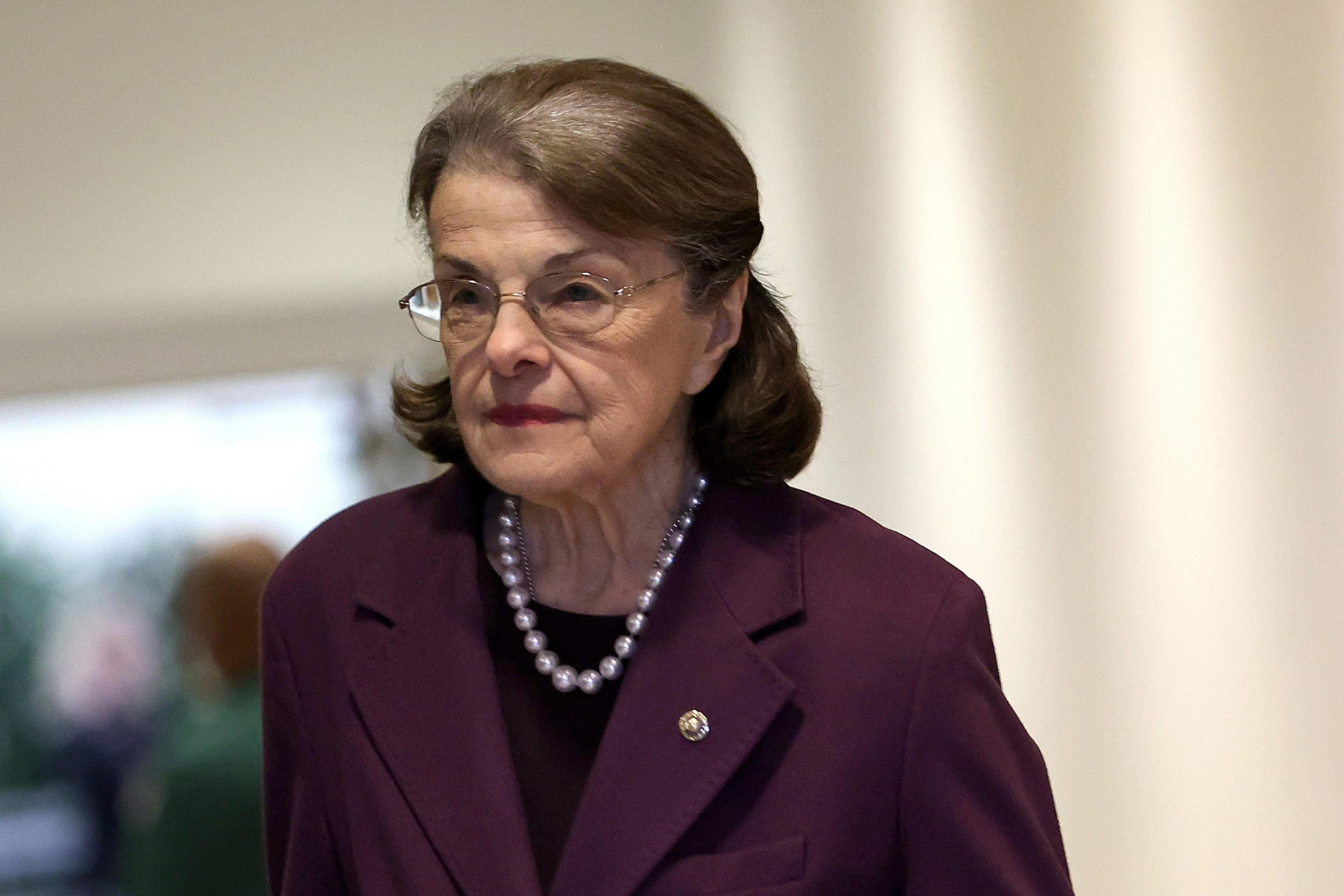 An older woman with short brown hair wears glasses, a dark purple blazer with a pin, a black top, and a pearl necklace, walking indoors with a neutral expression.