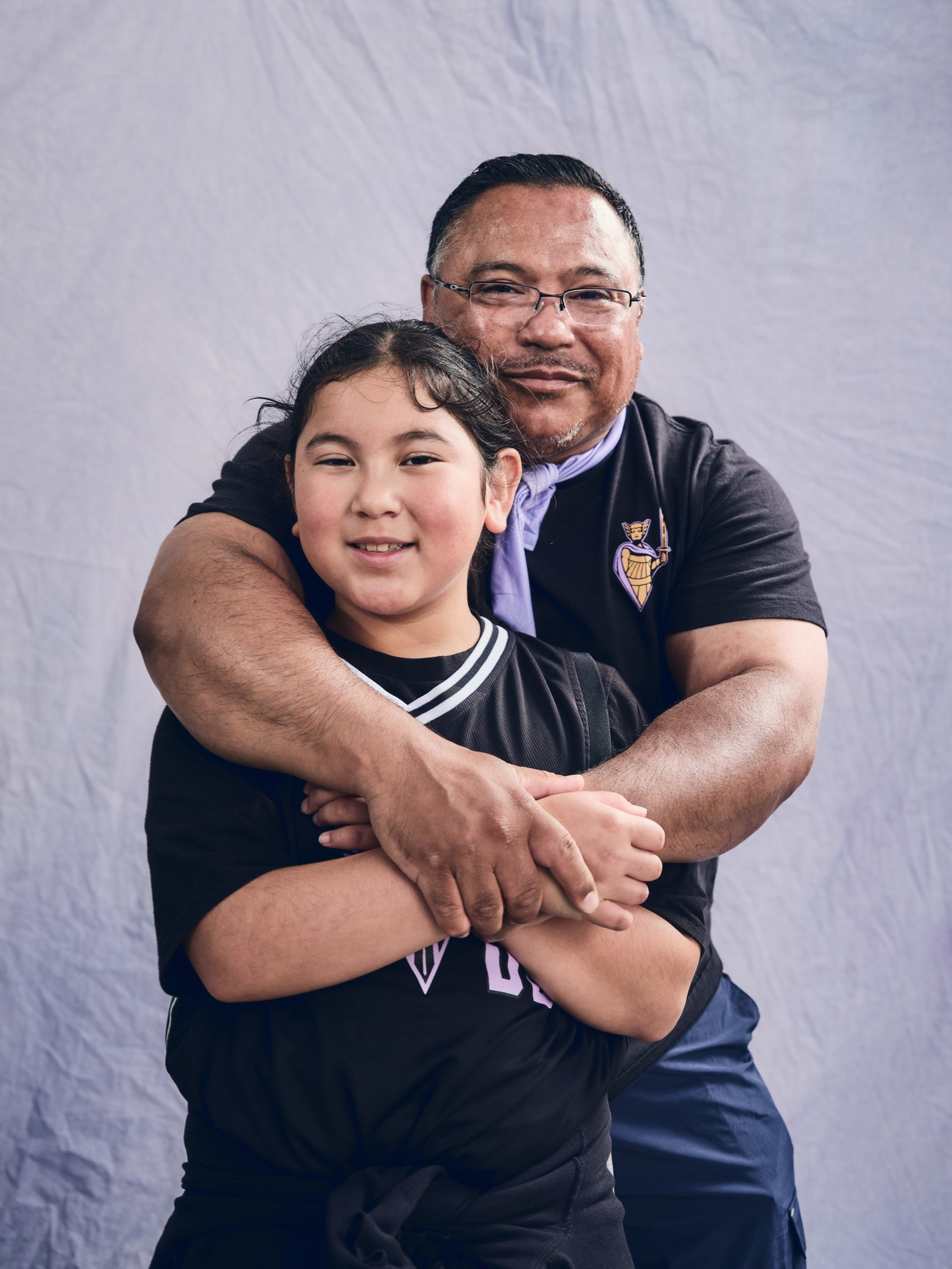 A man wearing glasses hugs a smiling girl from behind, both dressed in black; a light blue backdrop is behind them.