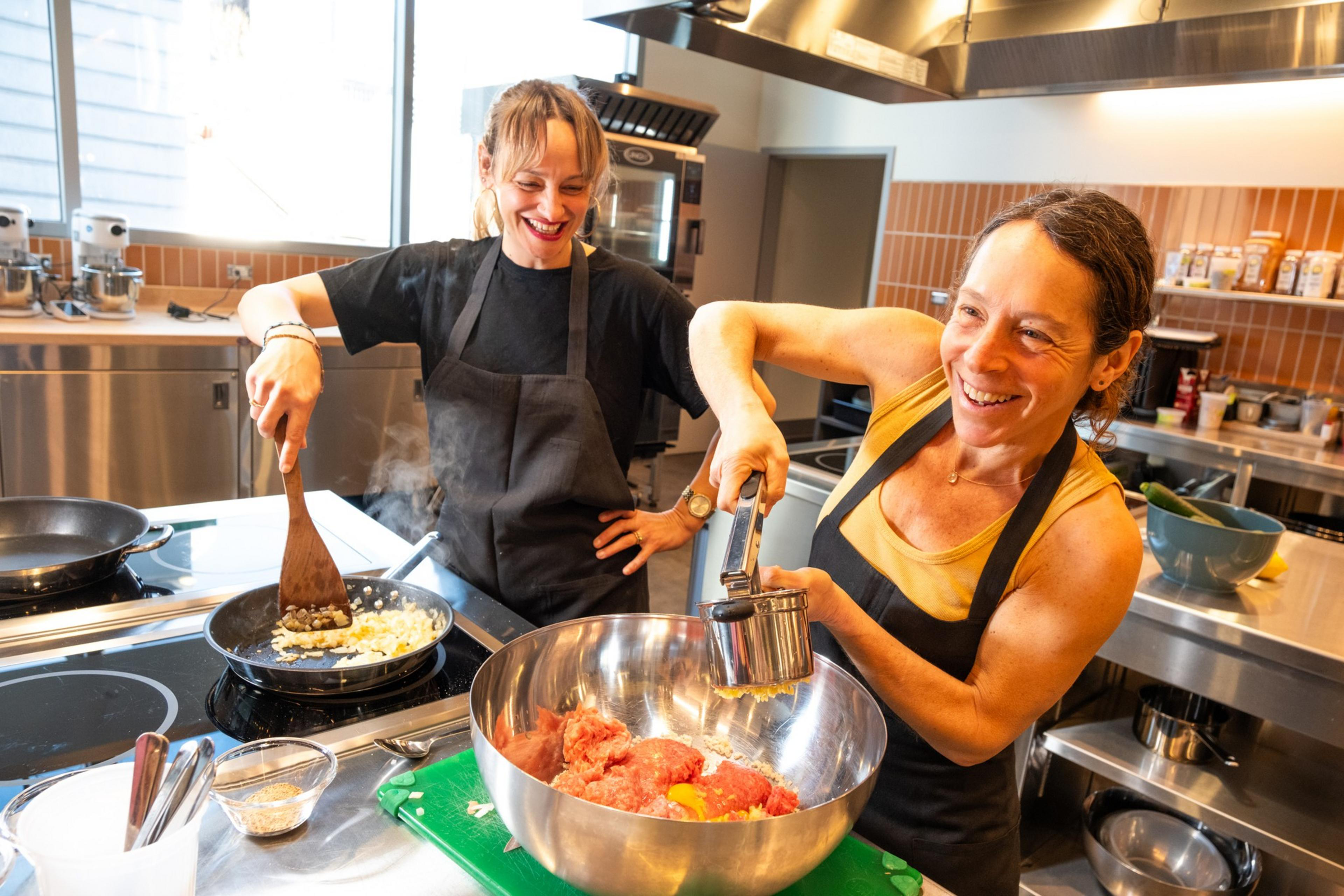 Two women are cooking in a kitchen, both wearing aprons. One is stirring onions in a pan, while the other is pressing garlic into a bowl with ground meat.