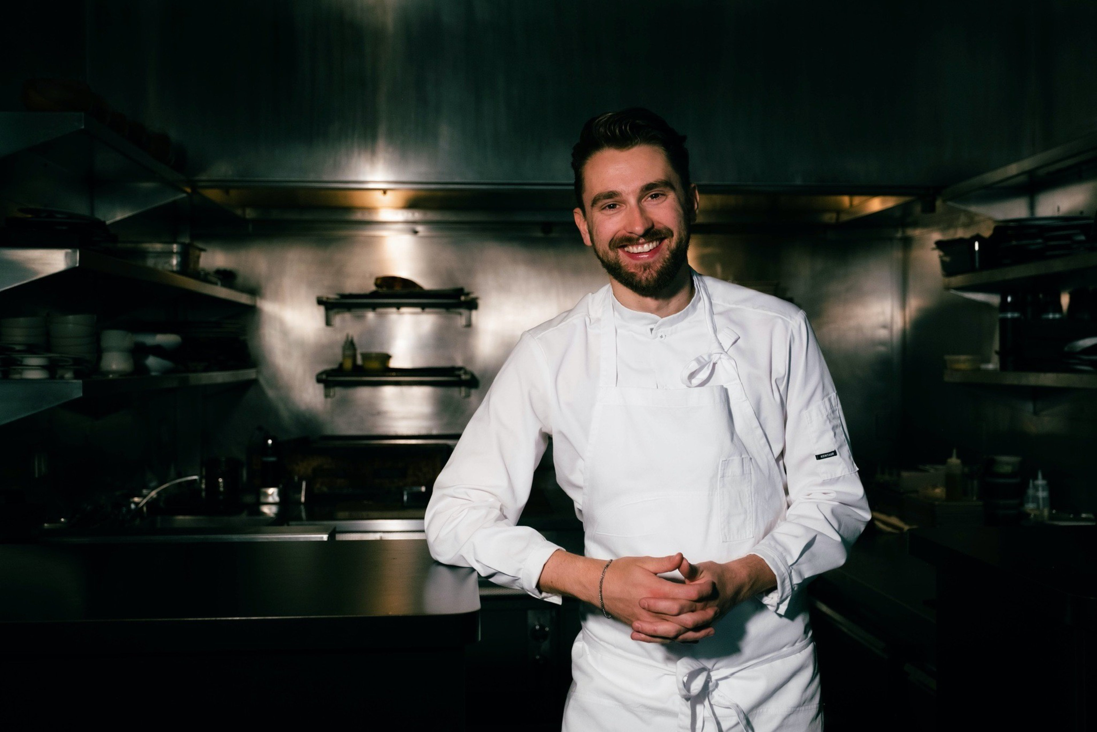 A smiling chef in a white uniform stands in a stainless-steel kitchen, hands clasped, with shelves and kitchen equipment in the background.