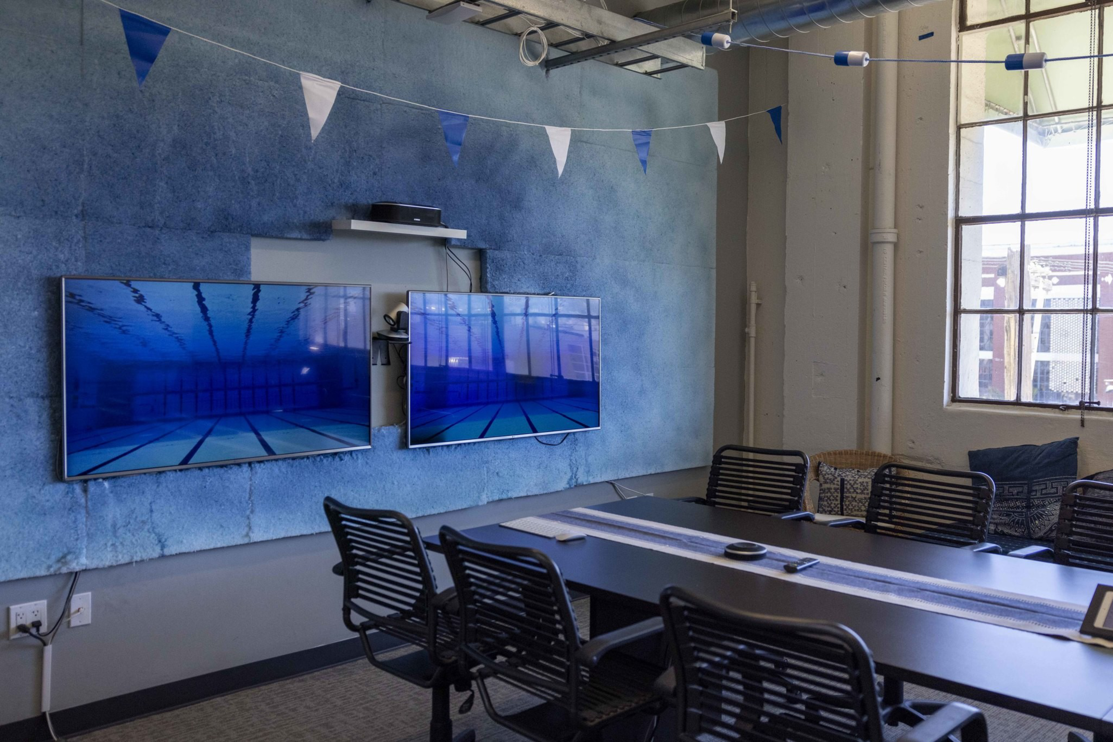 A conference room with six black chairs around a rectangular table, two wall-mounted screens displaying underwater pool scenes, and blue-white bunting overhead.