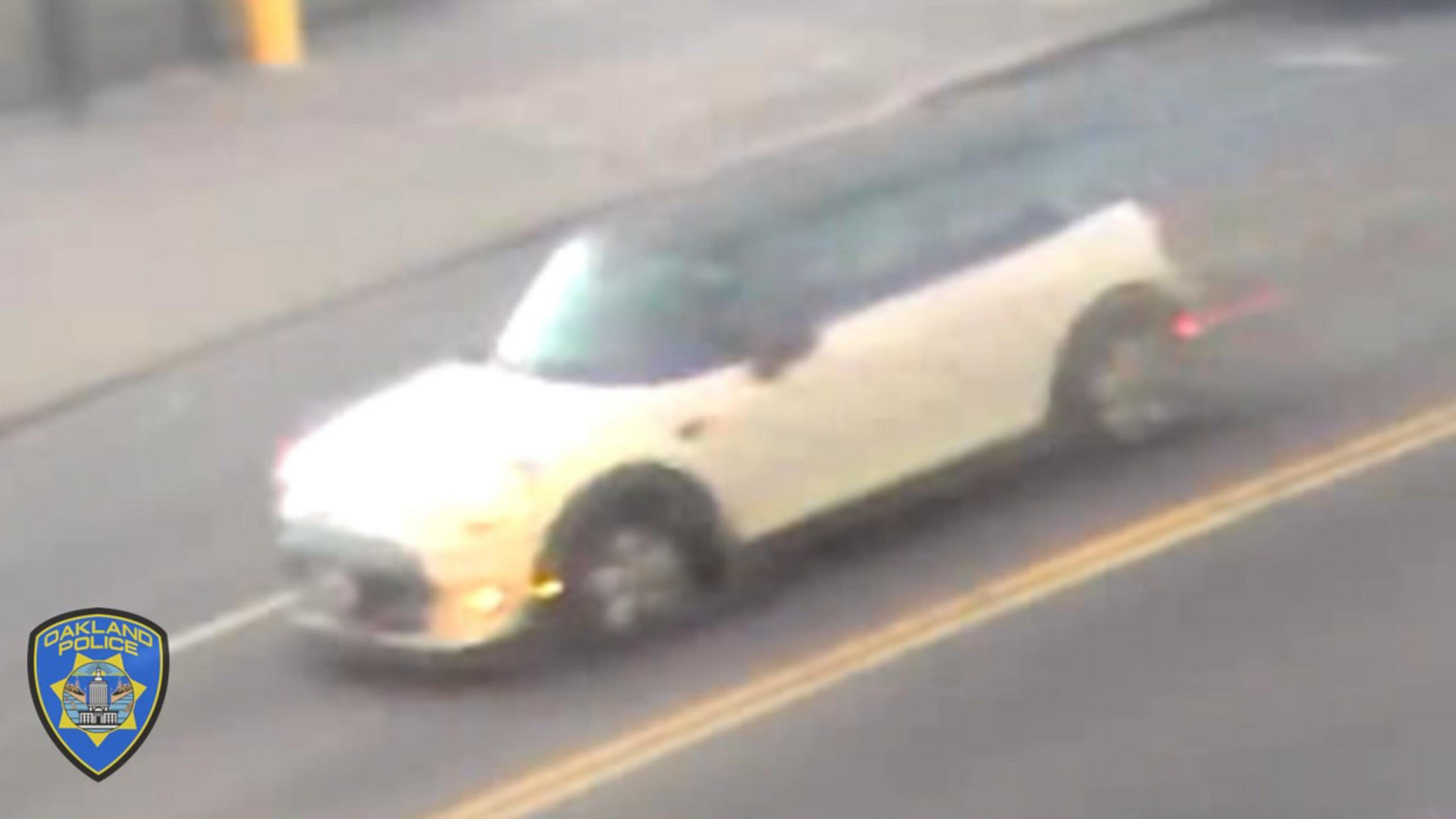 A beige Mini Cooper with a black roof is driving on a city street, captured in motion, with the Oakland Police badge visible in the corner.