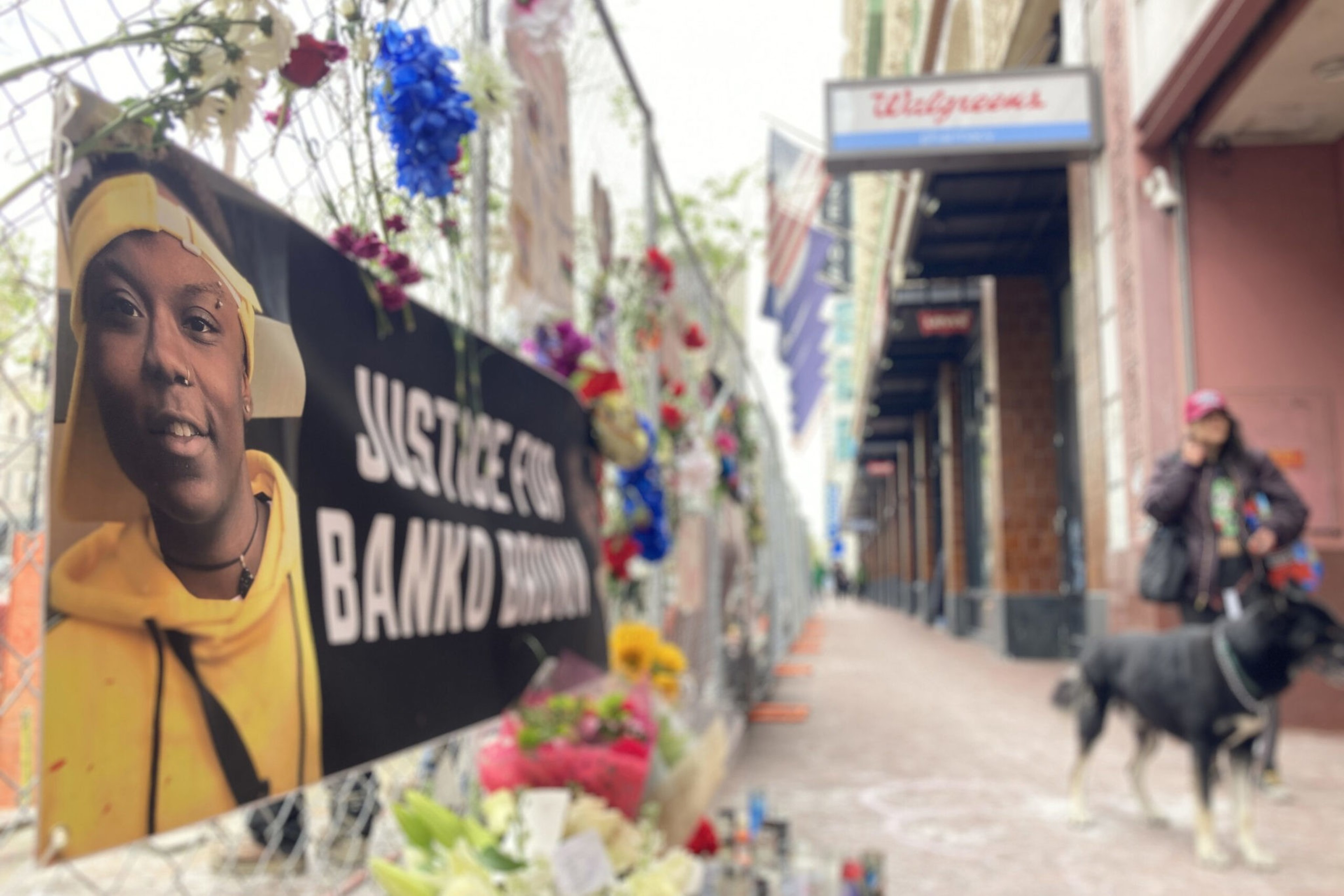 A memorial with flowers and a sign reading "Justice for Banko Brown" hangs on a fence outside a Walgreens, with a person and dog blurred in the background.