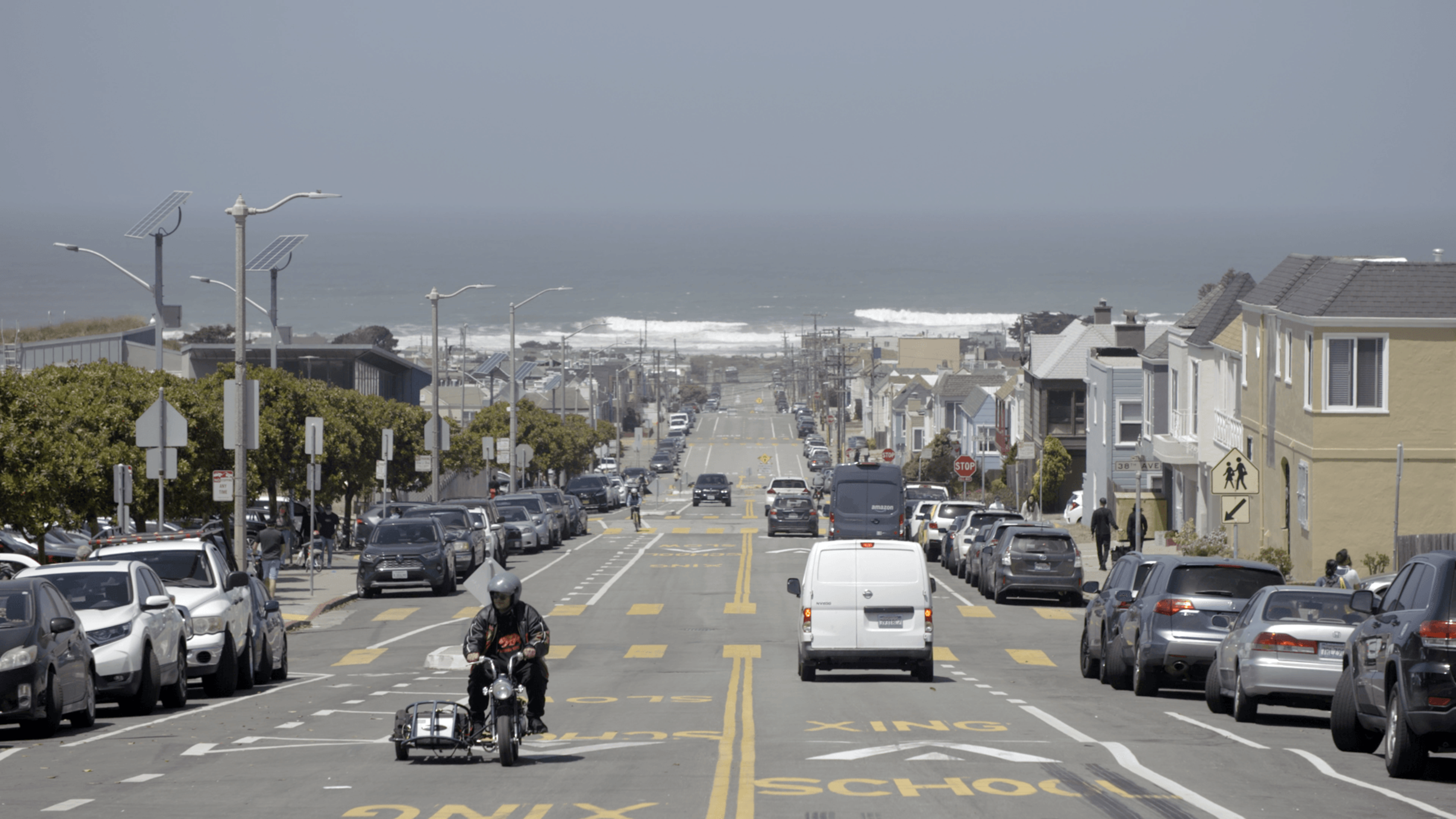 A wide street lined with parked cars on both sides slopes gently toward the ocean, with a motorcyclist, a white van, and a few pedestrians visible.