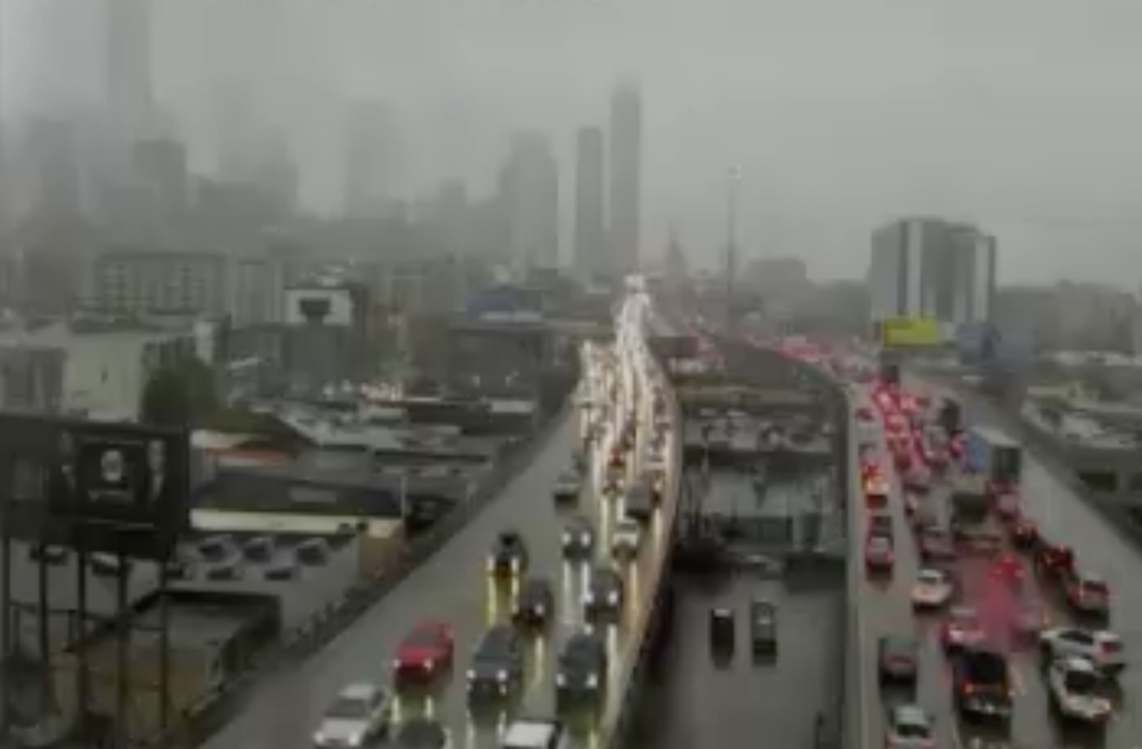 The image shows a busy, rain-soaked city highway with heavy traffic. Cars have their headlights on, and skyscrapers are barely visible through the fog.