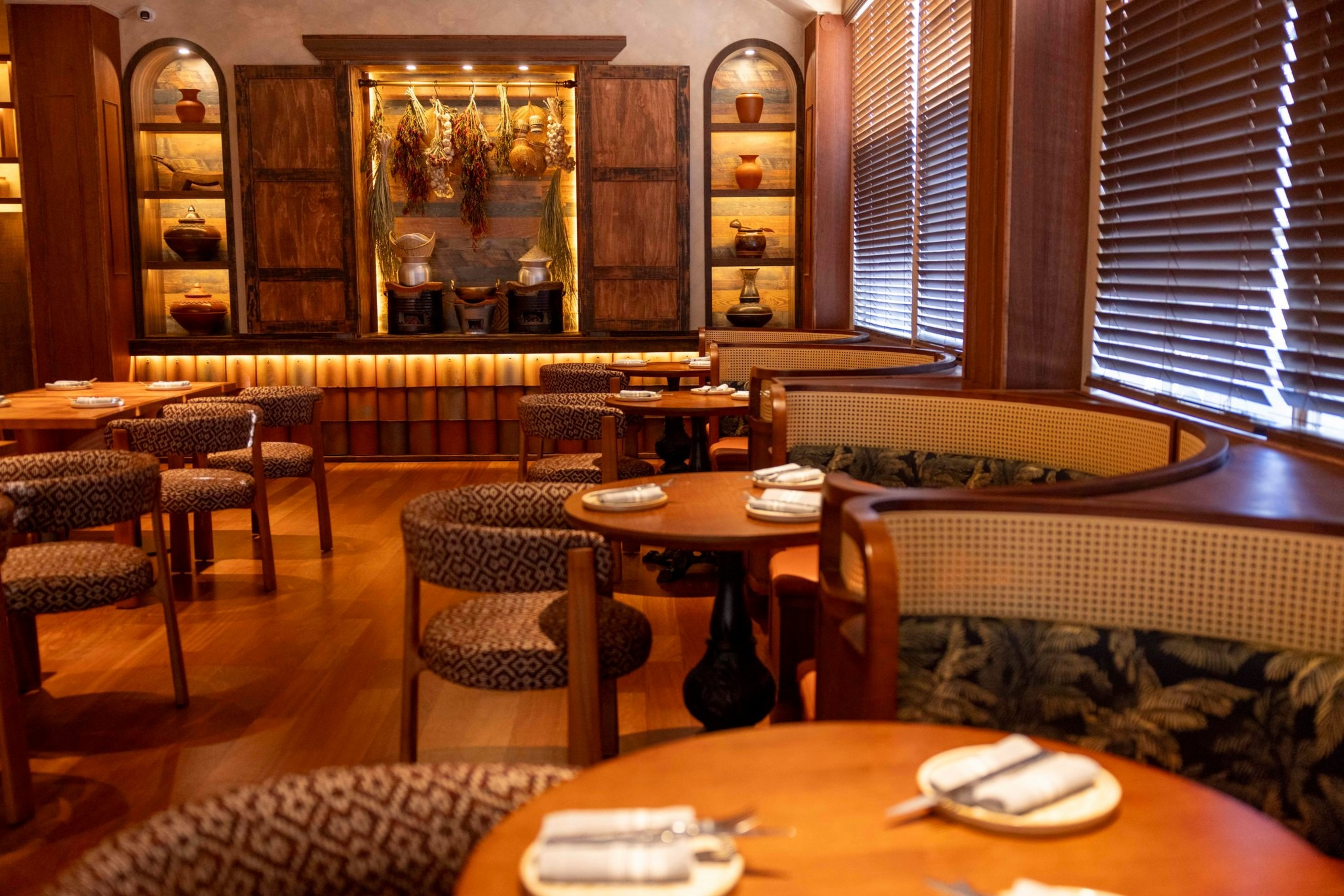 Cozy wooden restaurant interior with round tables, patterned chairs, curved booths, warm lighting, blinds on windows, and decorative hanging dried herbs.