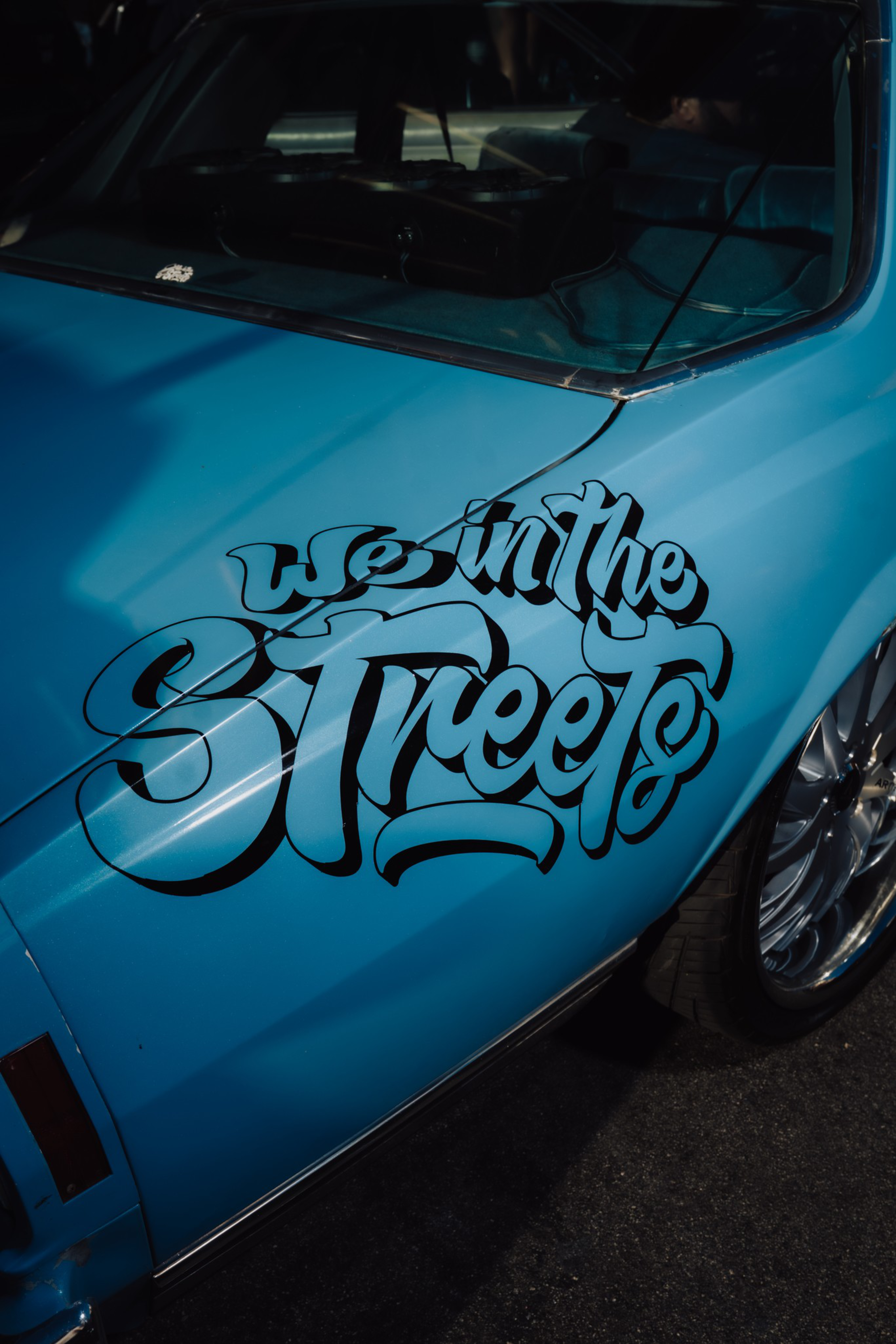 A blue car door with shiny chrome rims features bold, stylized black lettering that reads “we in the streets.”