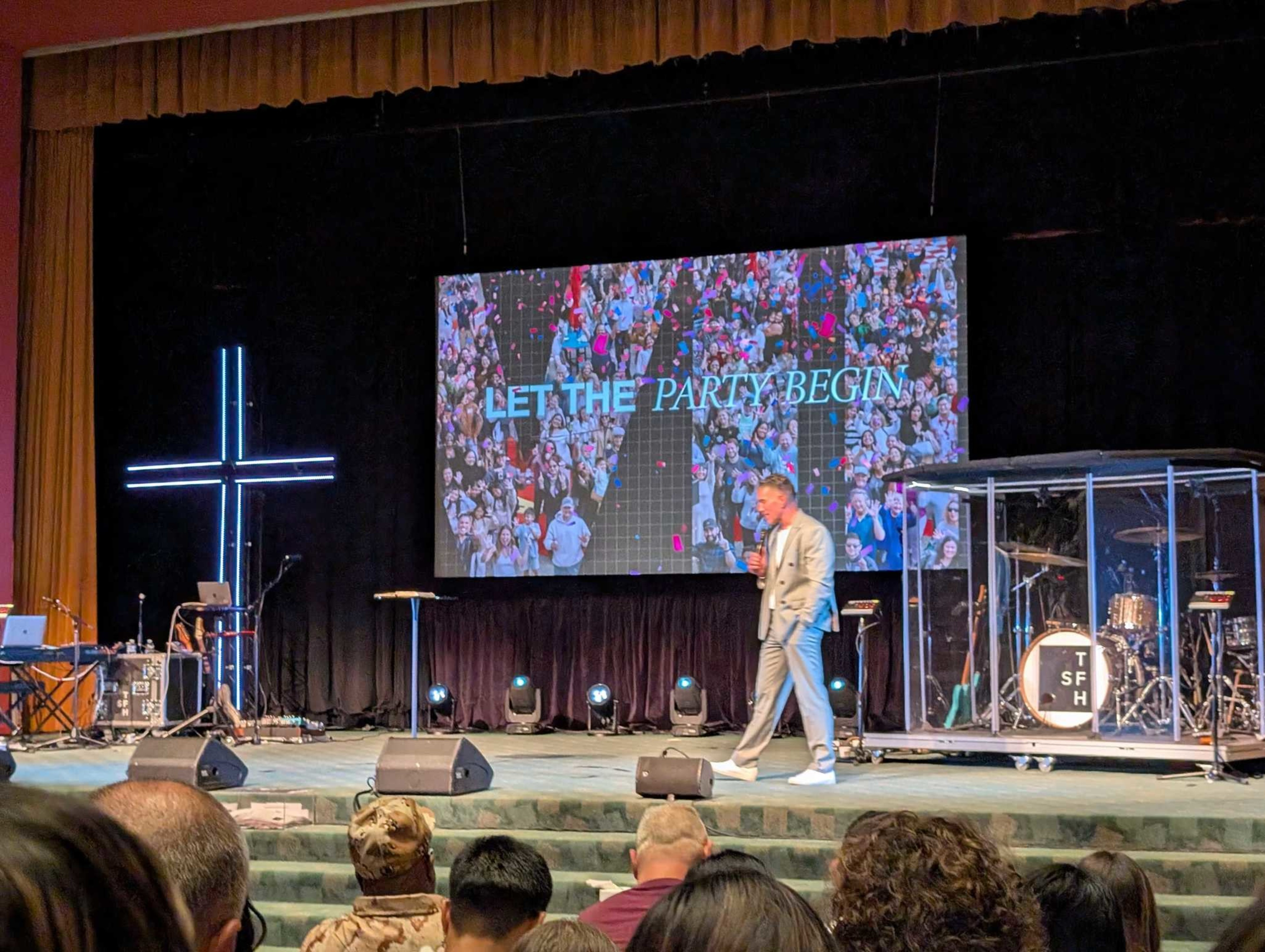 A man in a light suit speaks on stage in front of a large screen reading “LET THE PARTY BEGIN” with a crowd image, next to a glowing neon cross and drum set.
