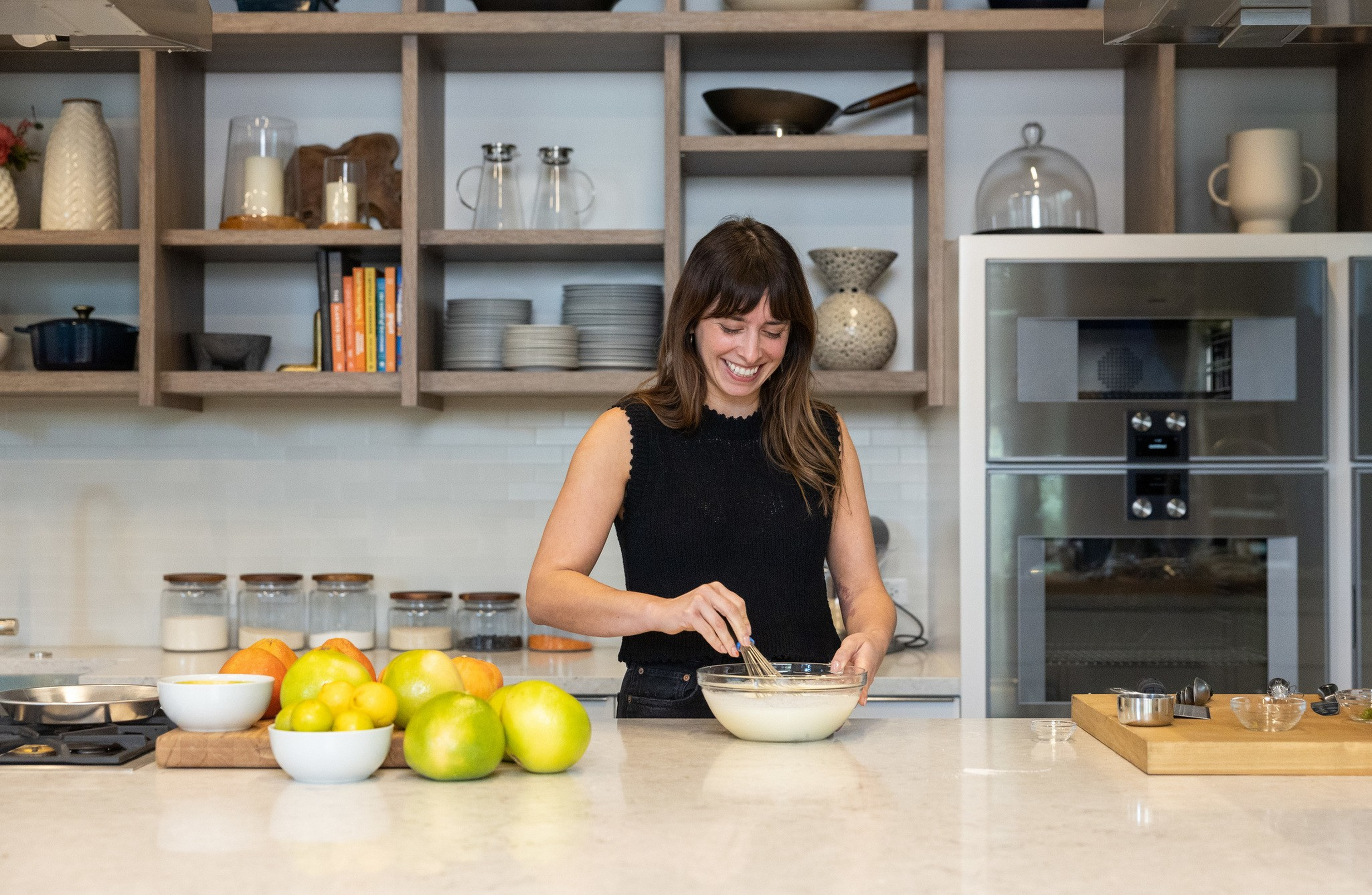 A woman in a black sleeveless top is whisking batter in a glass bowl on a kitchen counter with fruit and utensils nearby.
