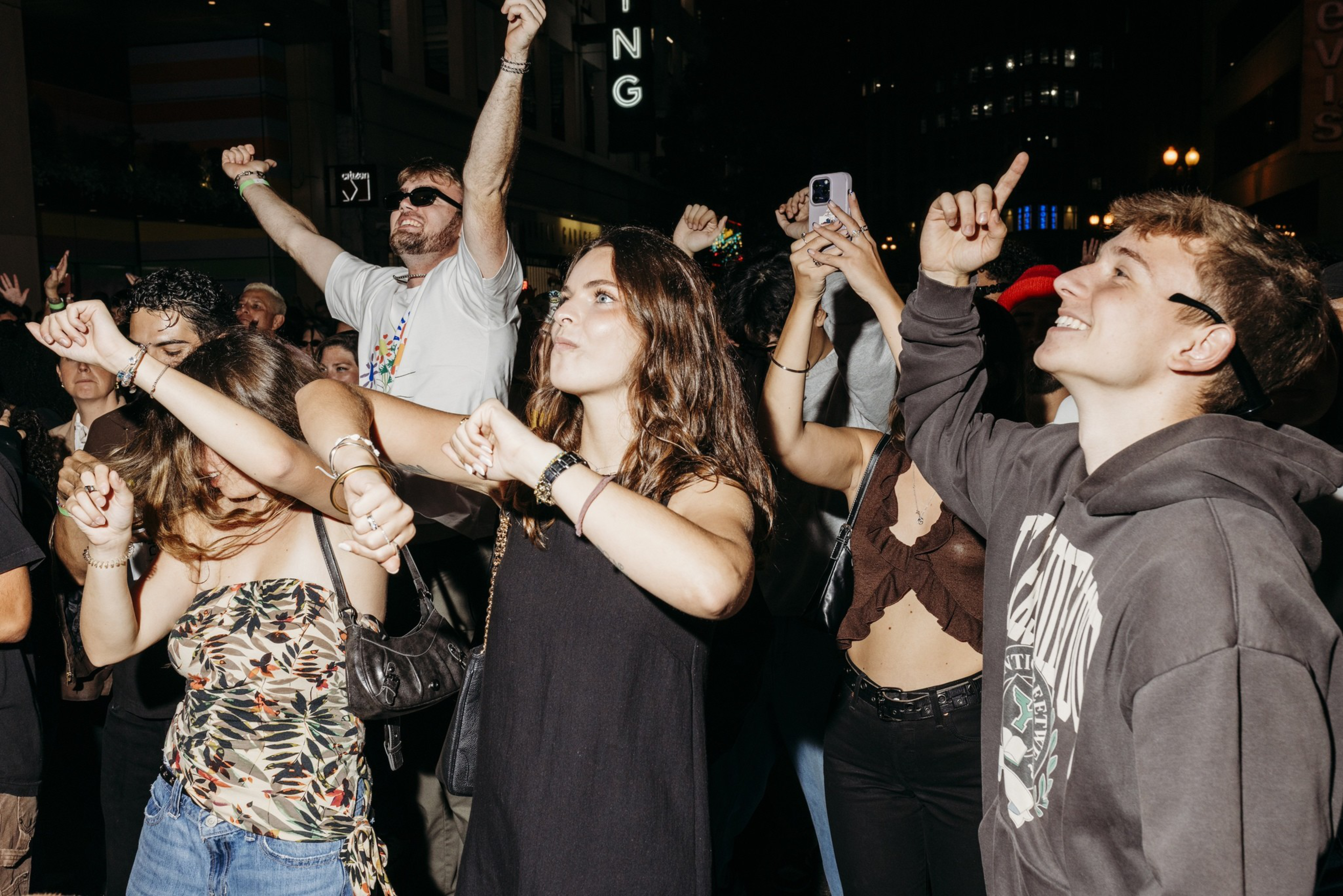 A group of young people dancing and smiling outdoors at night, some with raised arms, and one person holding a phone to record.