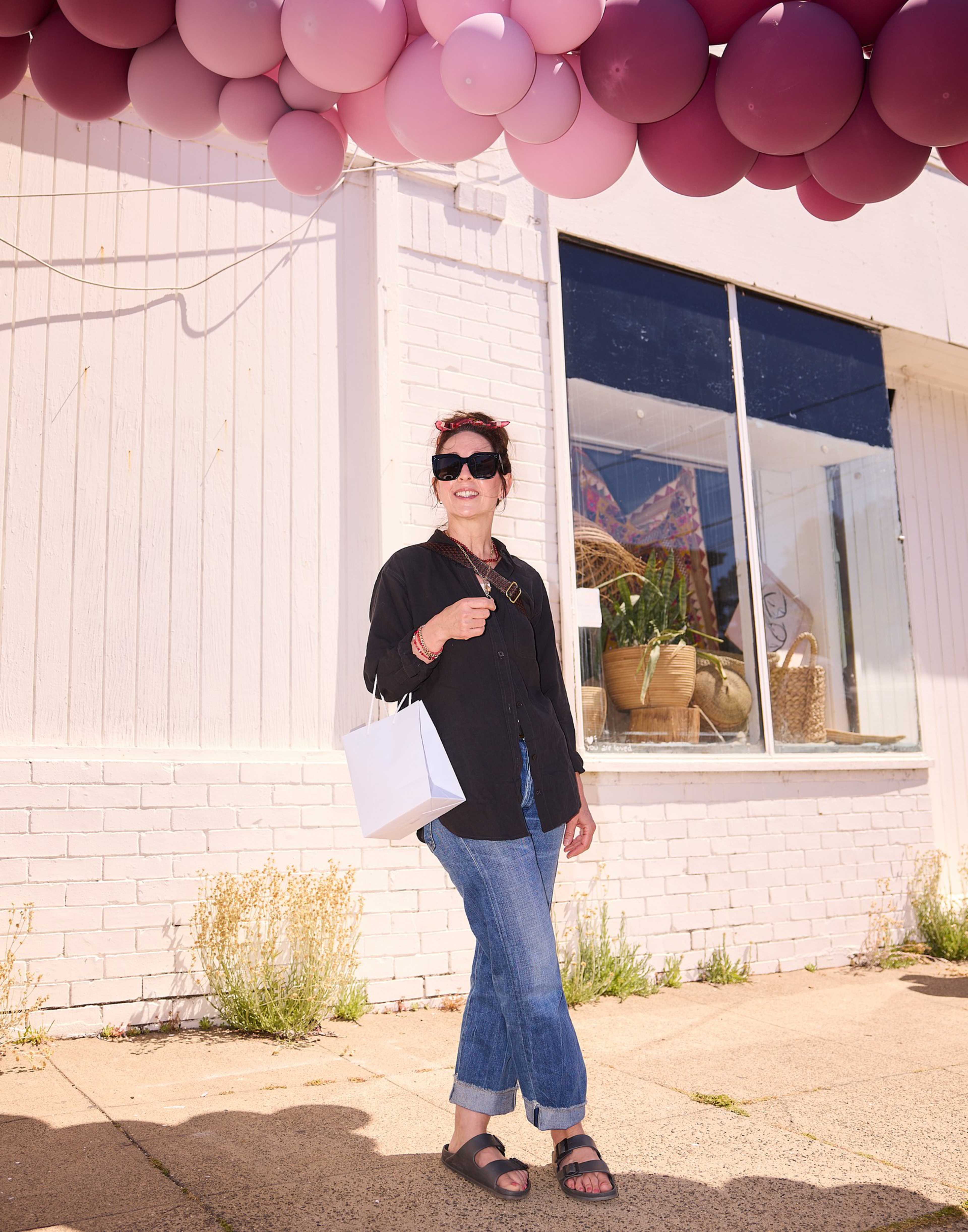 A woman wearing sunglasses, a black shirt, blue jeans, and sandals stands outside a white building under pink balloons, holding a white shopping bag.