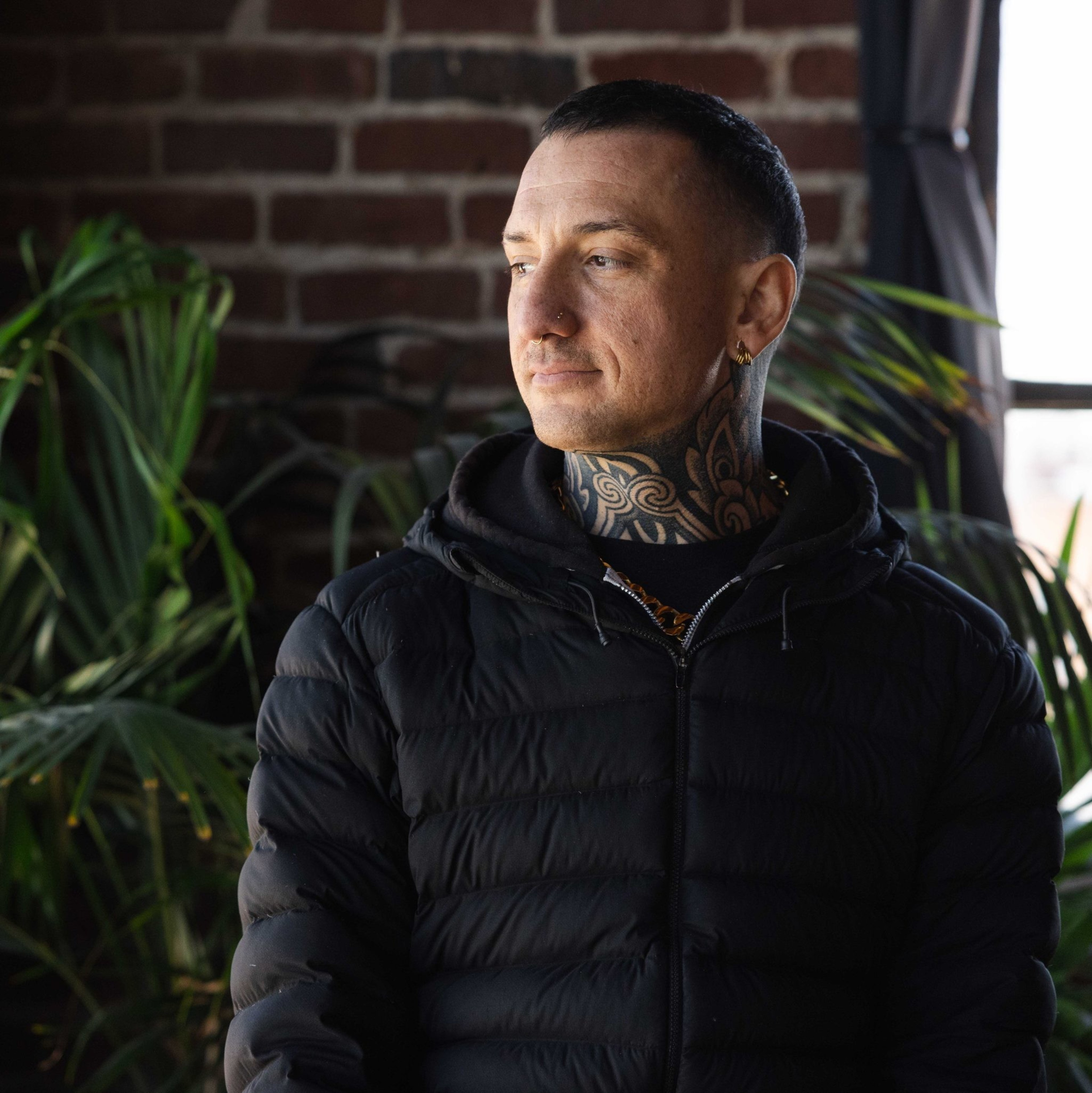 A person with a tattooed neck, wearing a black puffer jacket, looks thoughtfully to the side. They stand indoors near brick walls and green plants.