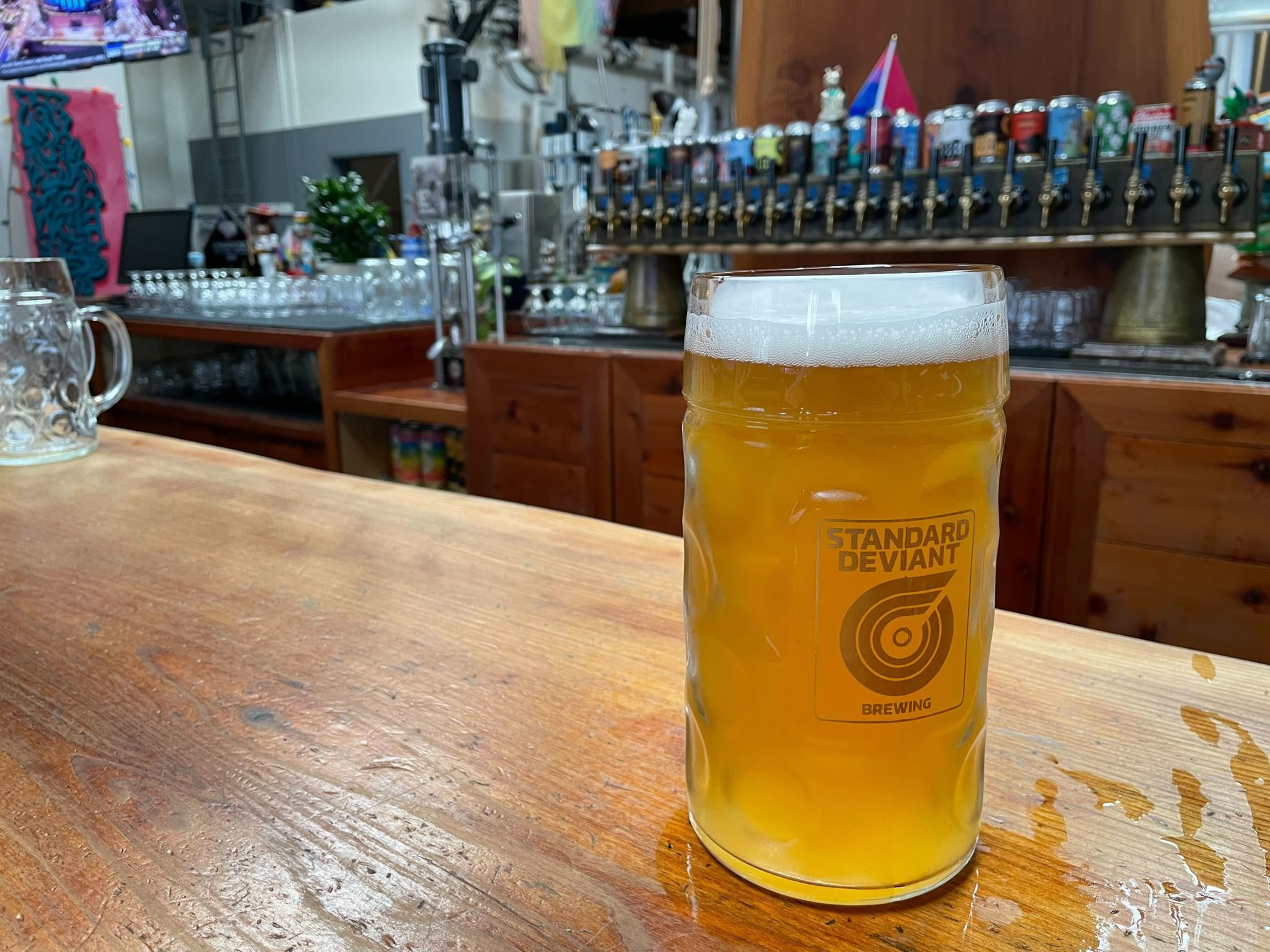A filled beer stein from &quot;Standard Deviant Brewing&quot; sits on a wooden bar counter, with a backdrop of numerous tap handles and a TV screen above.