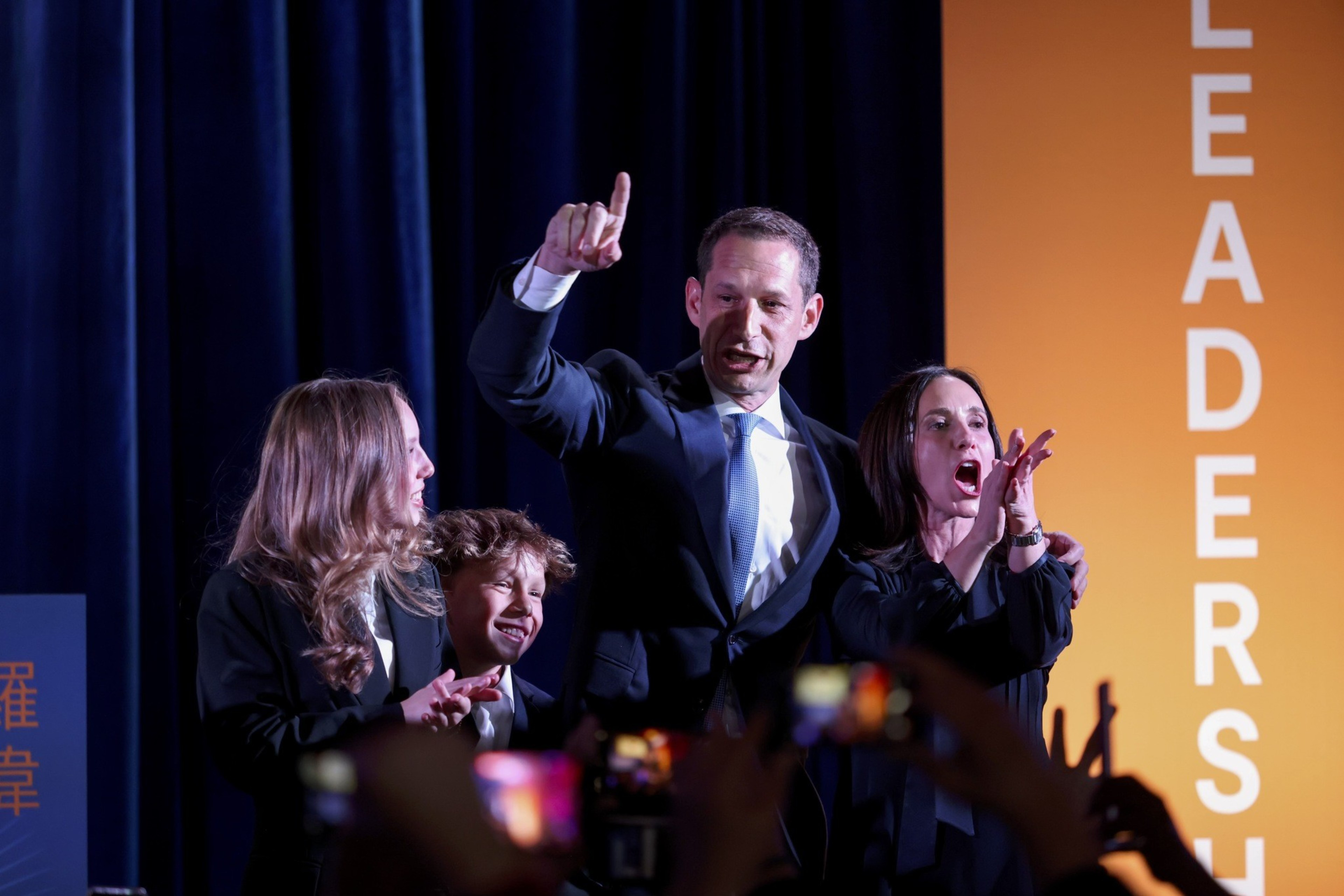 A man in a suit is raising his finger, flanked by two adults cheering and a child smiling, against a backdrop with "LEADERSHIP" in large letters.