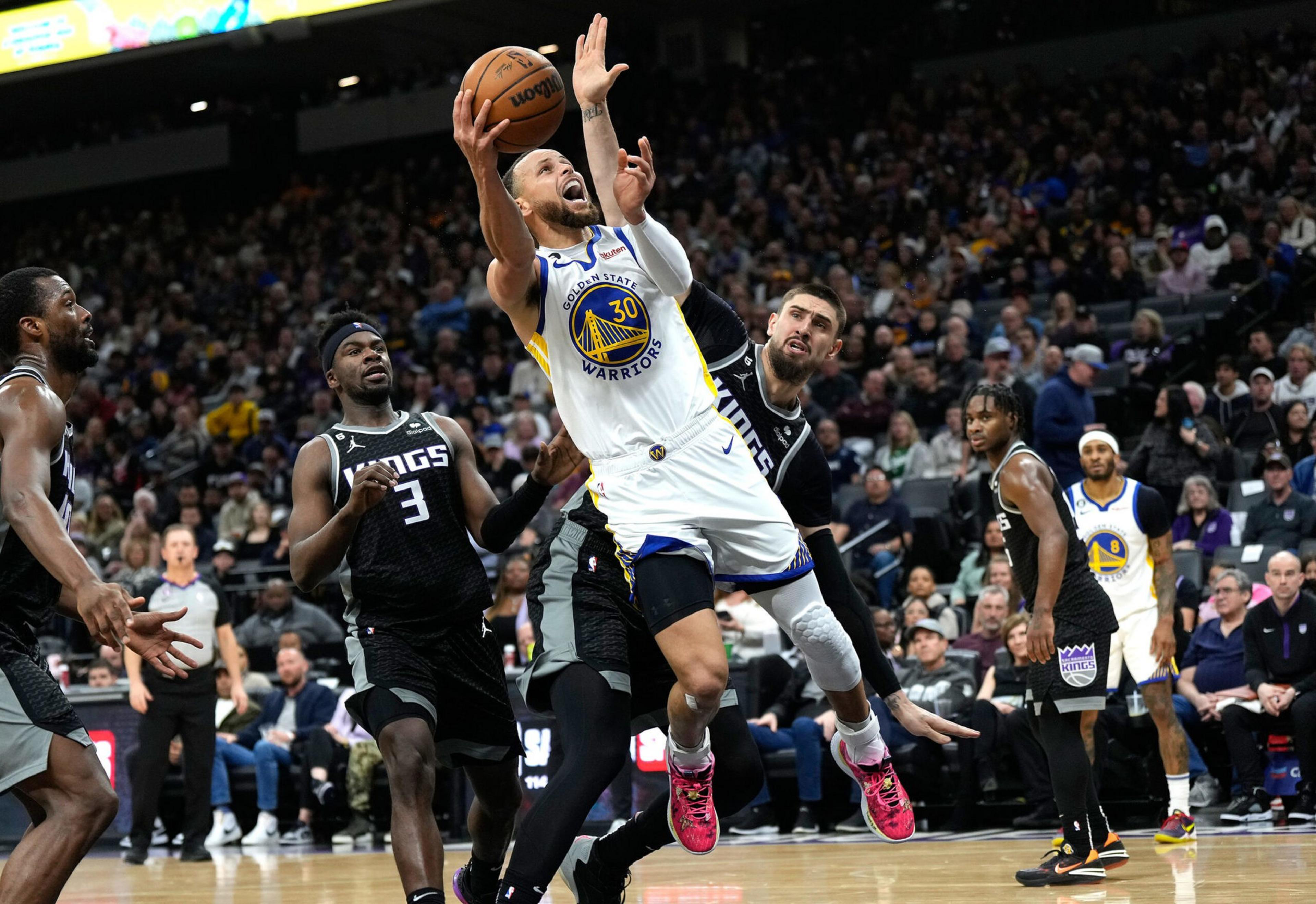 Stephen Curry, wearing a Golden State Warriors uniform, leaps toward the basket attempting a shot while surrounded by Sacramento Kings defenders.