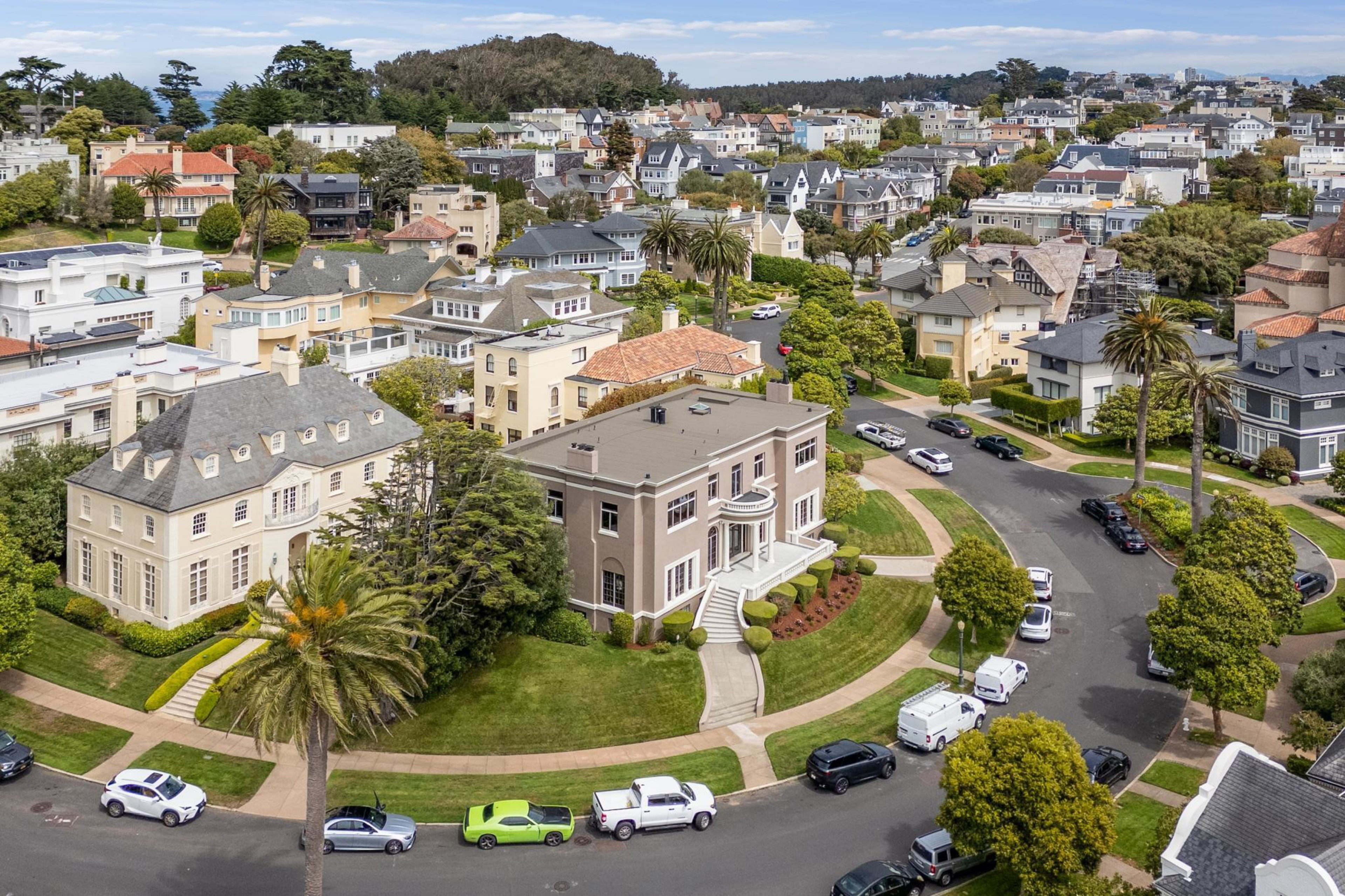 A neighborhood with large, elegant houses surrounded by green lawns and palm trees lines curved streets filled with parked cars.