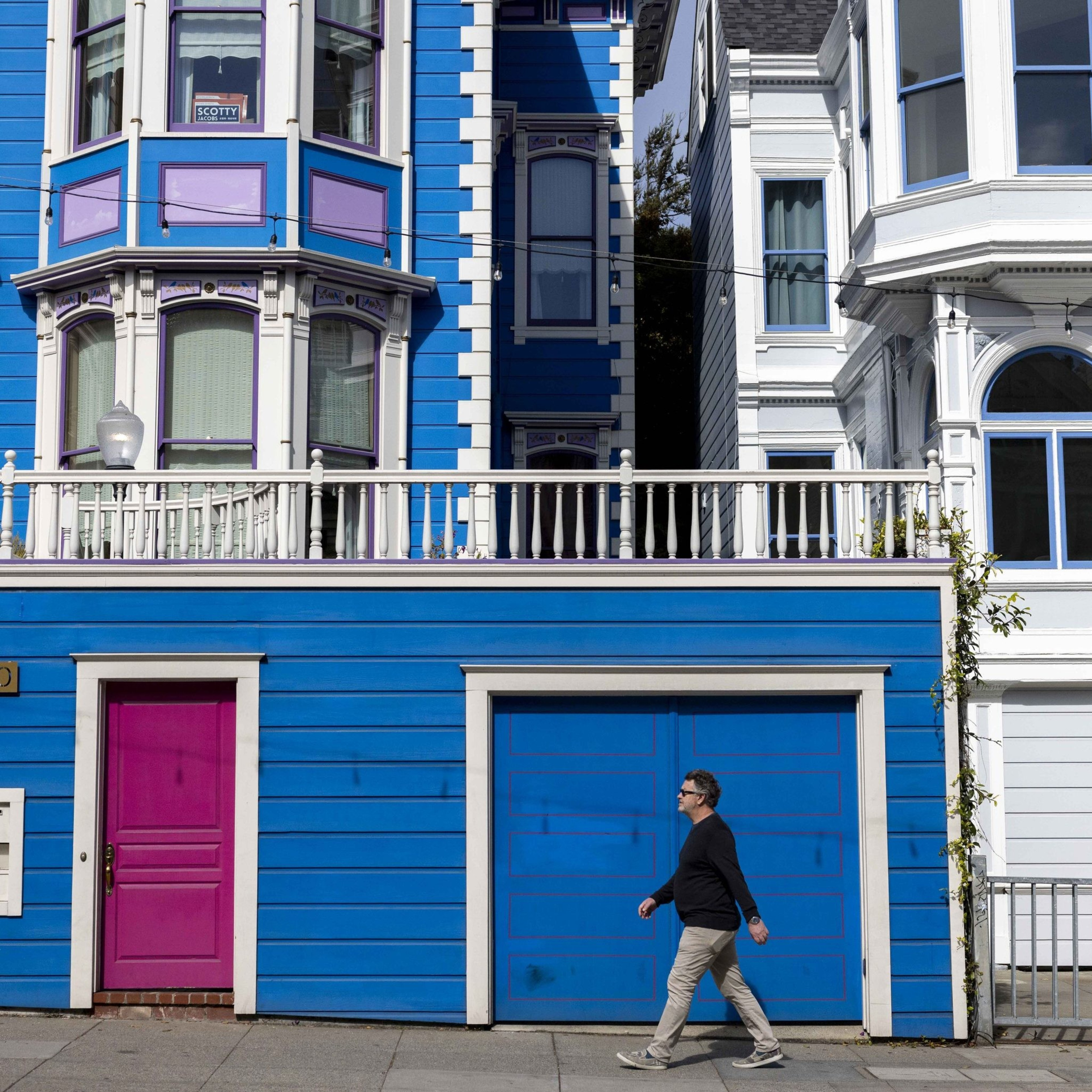 A man in a black jacket walks past a vibrant blue house with a pink door and blue garage, framed by white-trimmed windows and ornate detailing.
