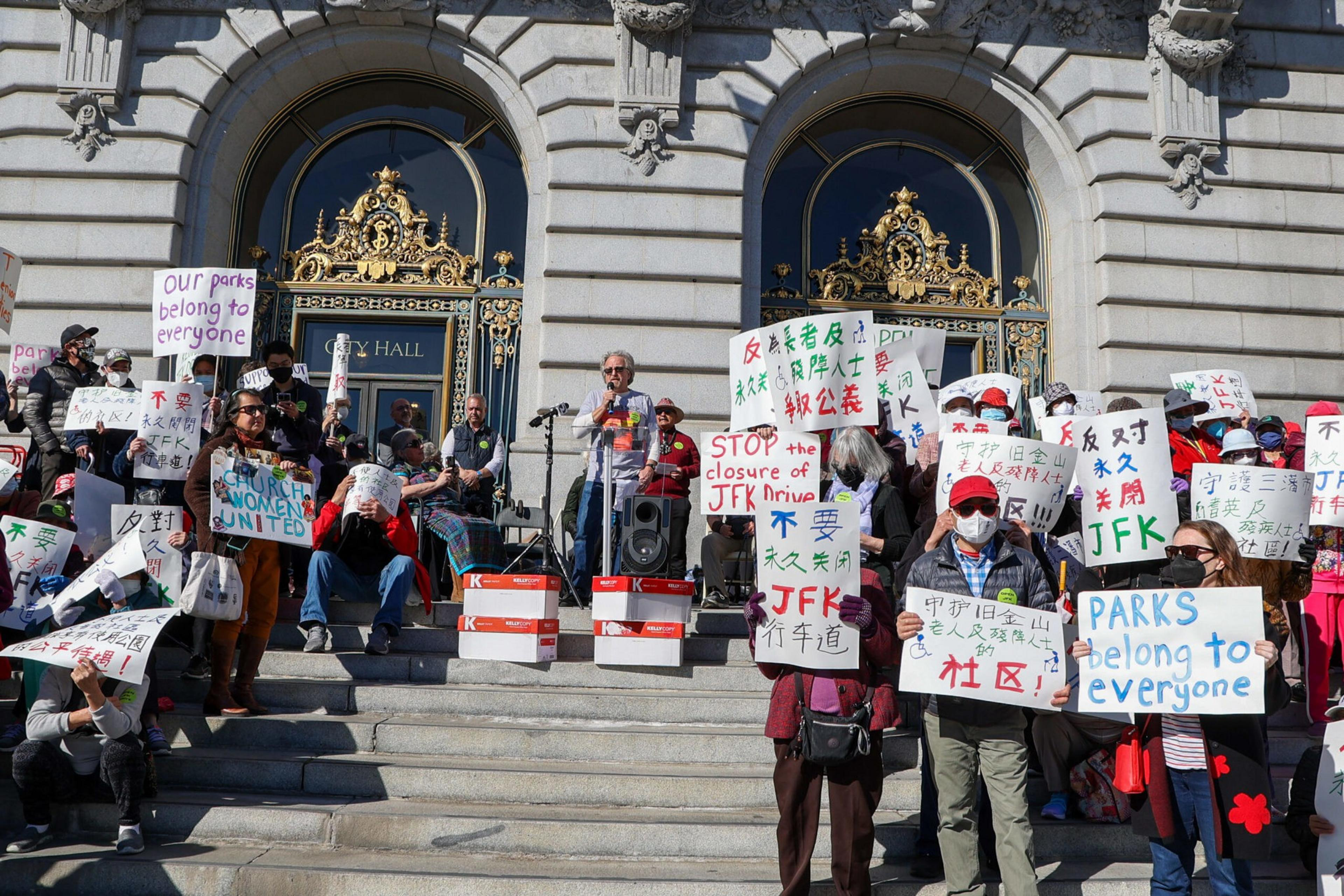 A crowd of masked protesters holds bilingual signs outside a grand building demanding to stop the closure of JFK Drive and that parks belong to everyone.