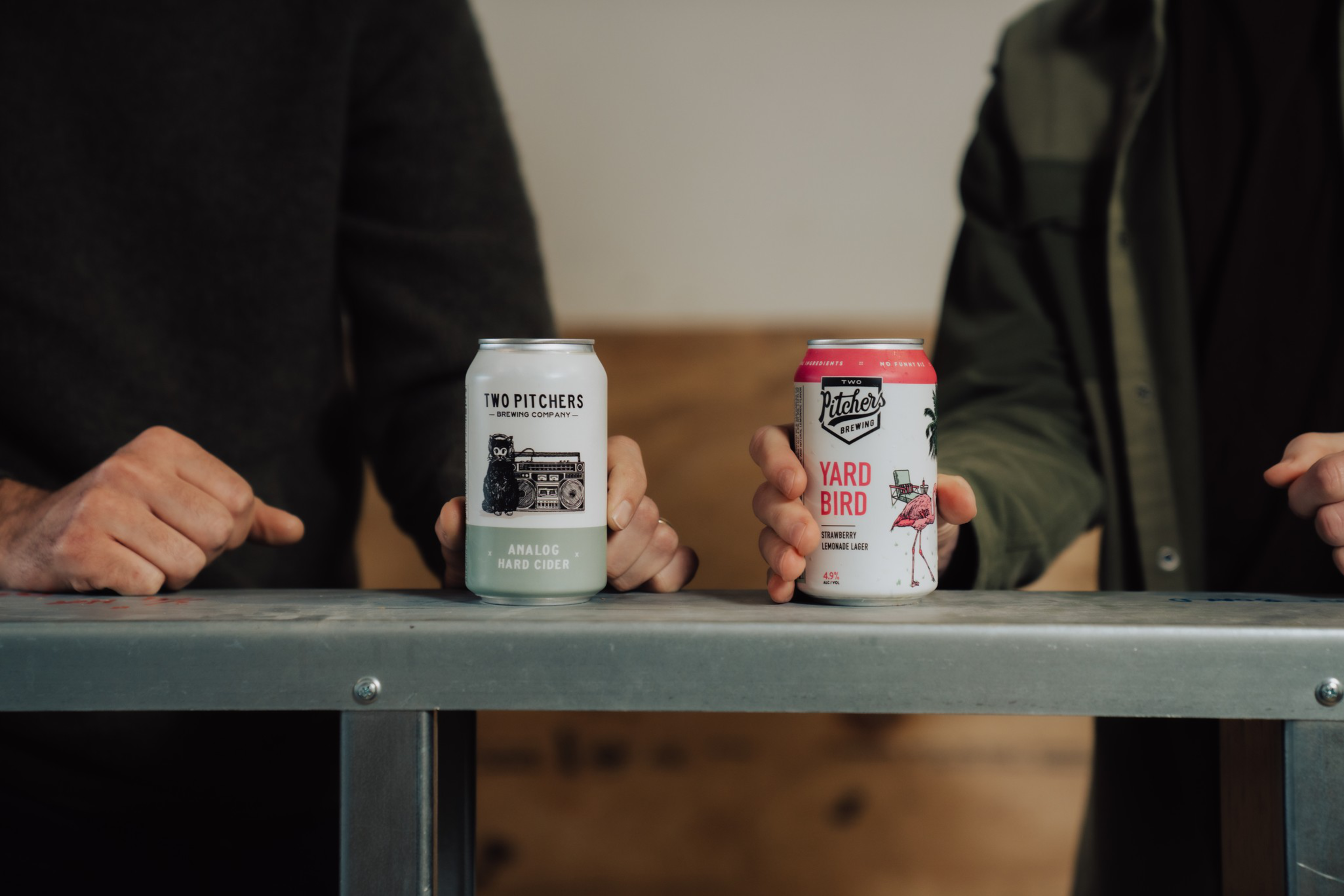 Two people each hold a can of craft beverage, one labeled “Two Pitchers Analog Hard Cider” and the other “Pitchers Yard Bird Strawberry Lemonade Lager.”