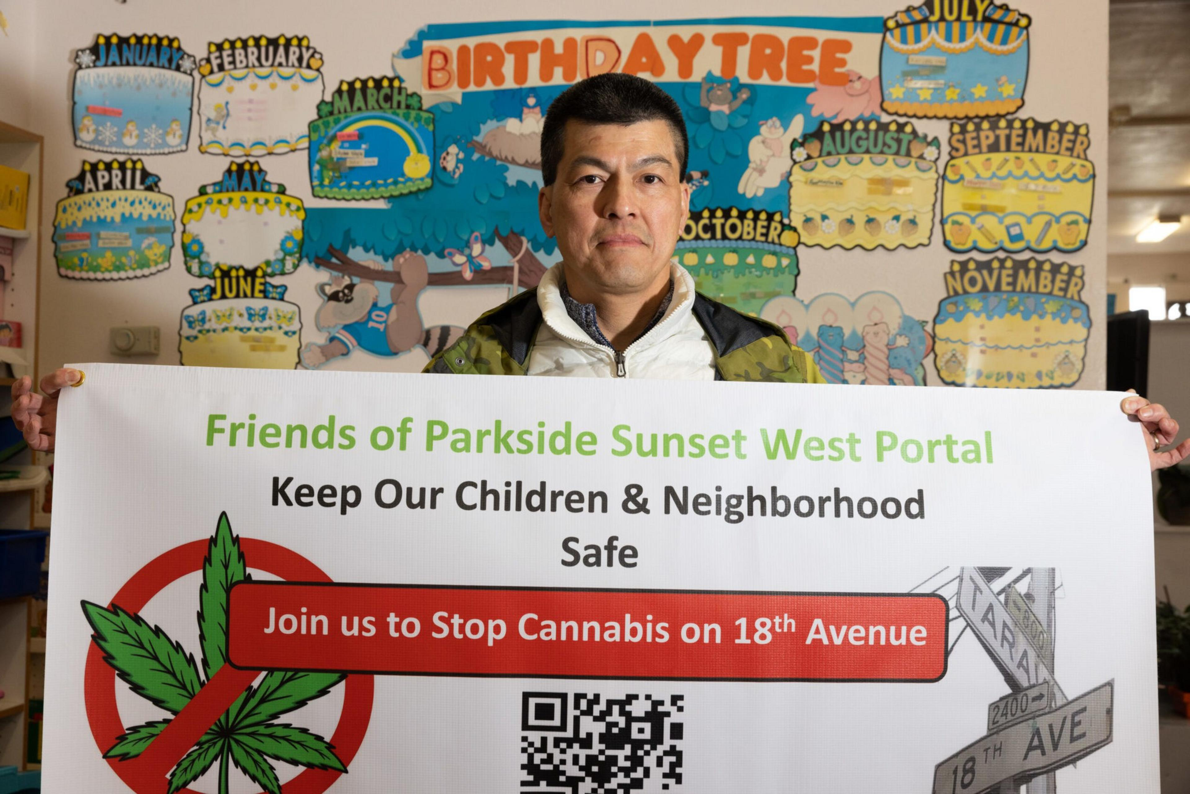 A man holds a sign against cannabis on 18th Avenue, with a cartoon birthday tree mural in the background showing months and decorations.