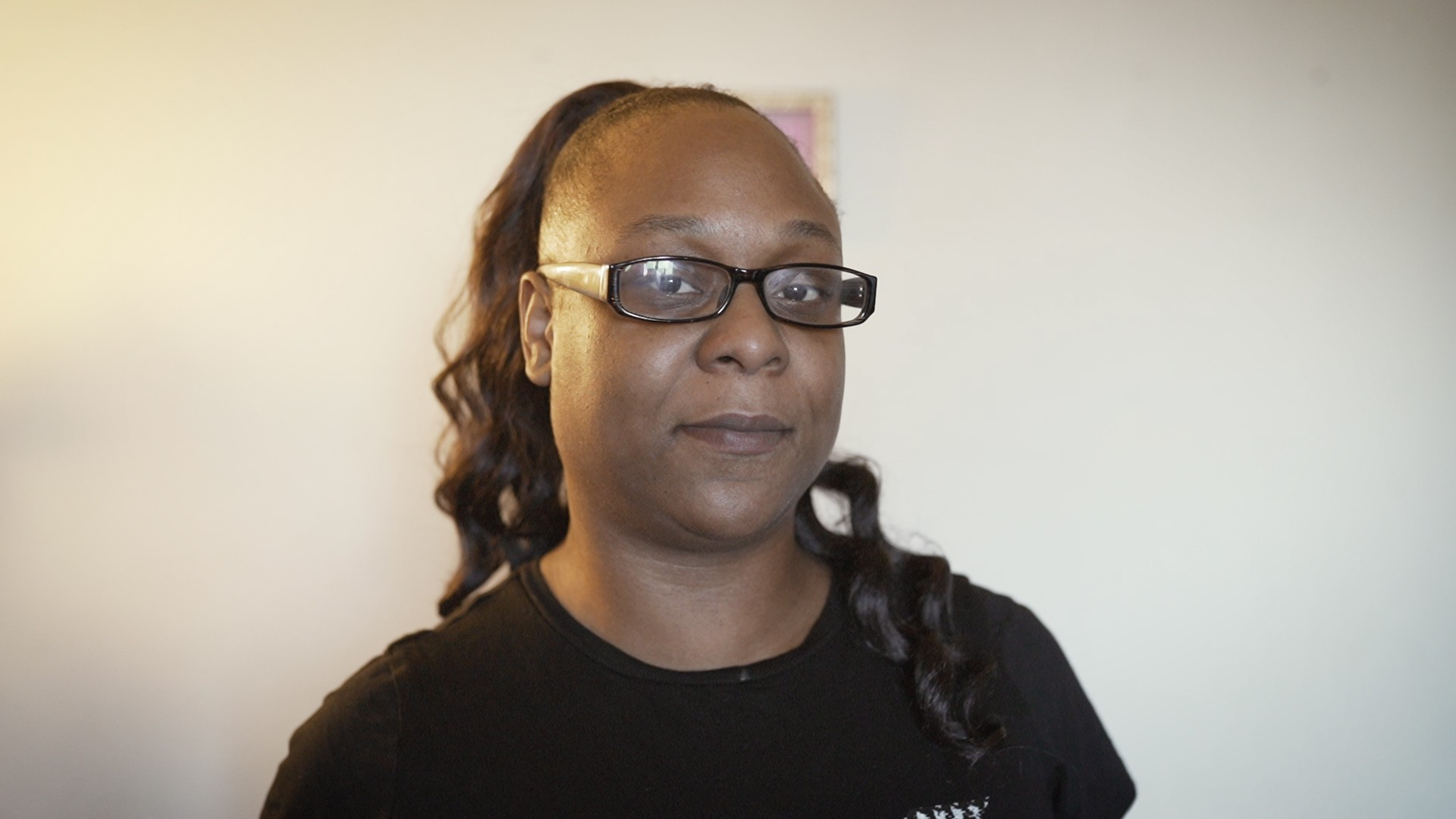 A woman with long, curly hair pulled back in a ponytail wears black rectangular glasses and a black shirt, standing against a plain light wall.