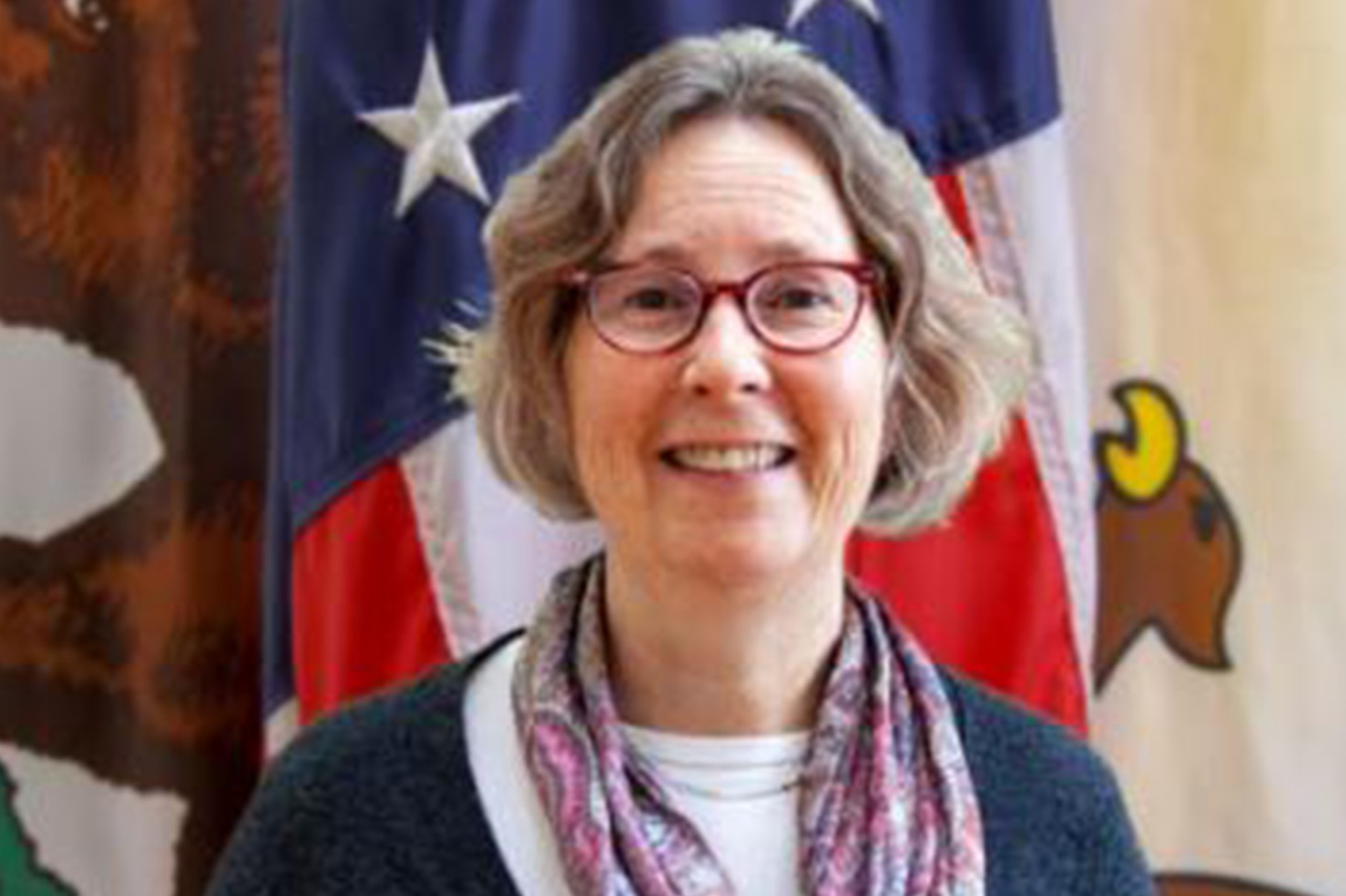 A smiling woman with short gray hair, red glasses, and a patterned scarf stands in front of an American flag and another flag with a bird emblem.