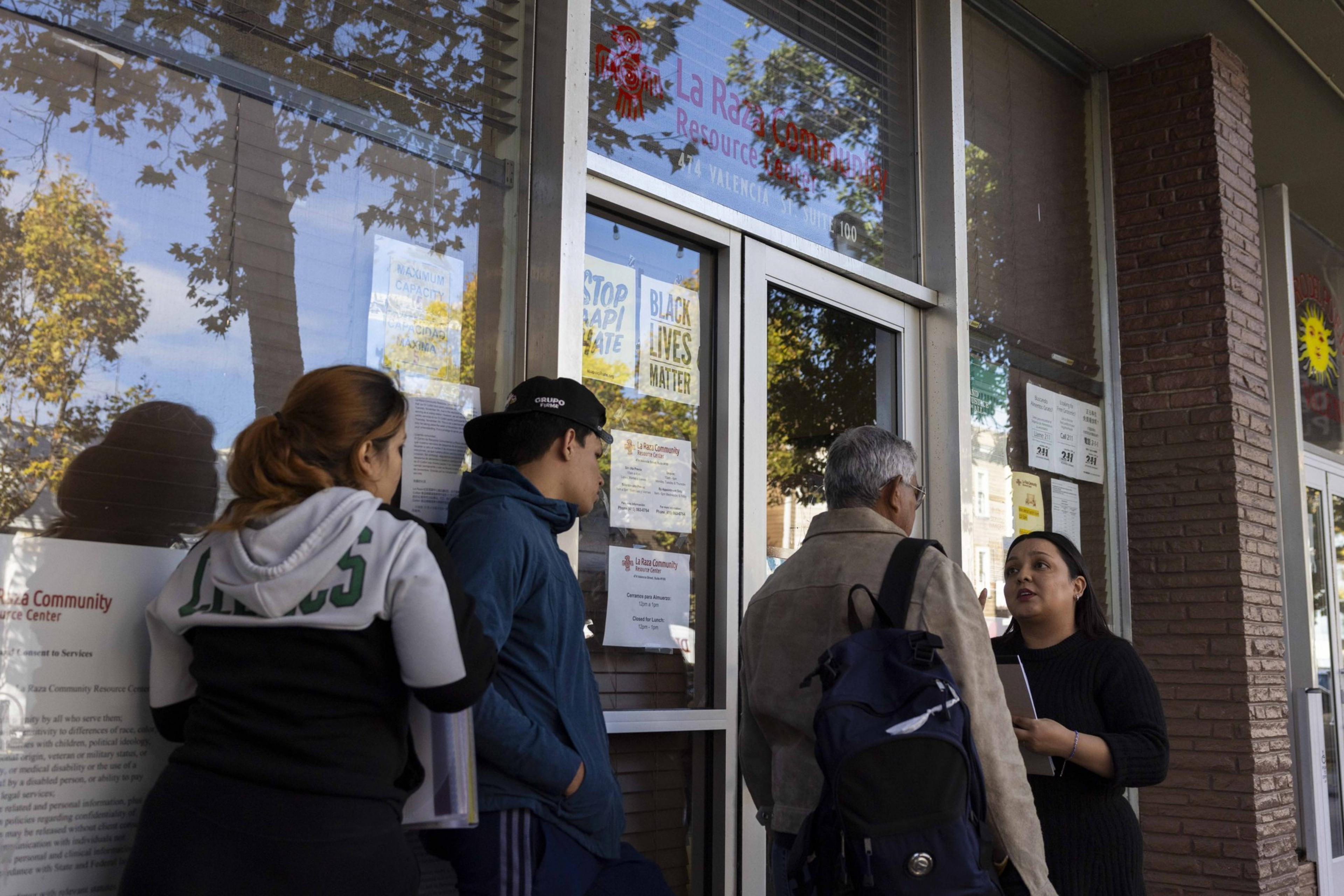 People are lined up outside a building with a glass entrance. A woman talks with a man holding a backpack, while others wait. Signs are visible on the window.