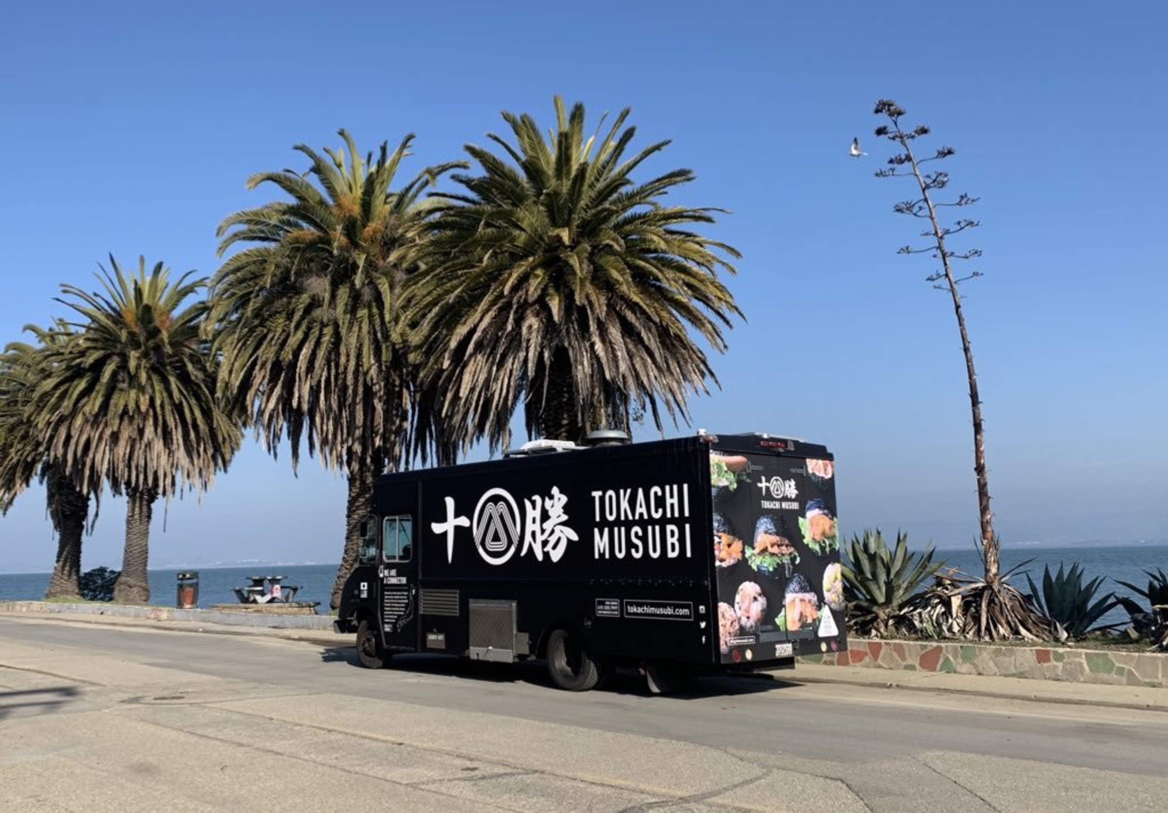 A black food truck with "TOKACHI MUSUBI" and Japanese characters is parked by a seaside road lined with palm trees under a clear blue sky.