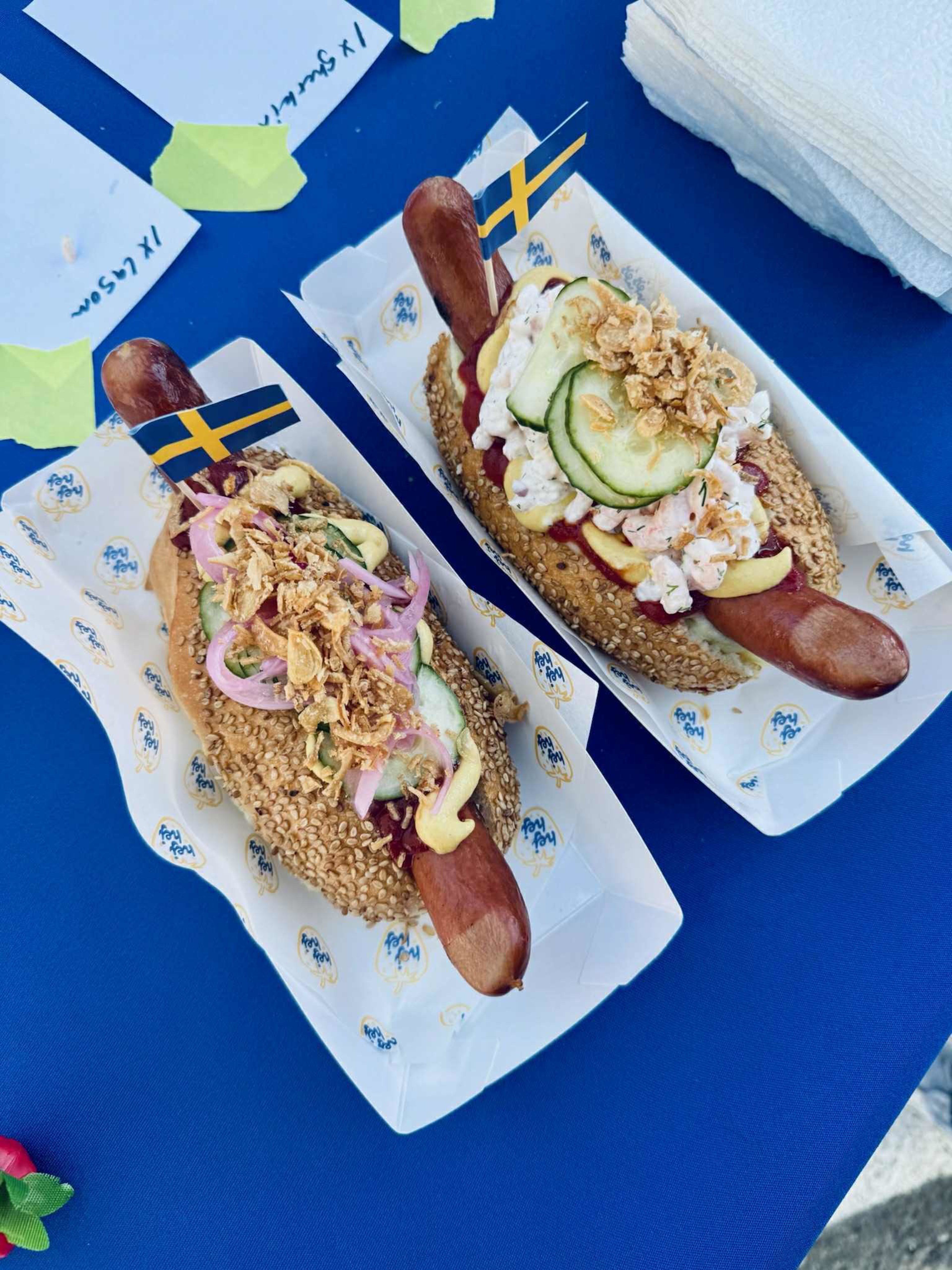 Two hot dogs in sesame seed buns topped with pickles, crispy fried onions, mustard, and creamy sauce, each with a small Swedish flag toothpick.