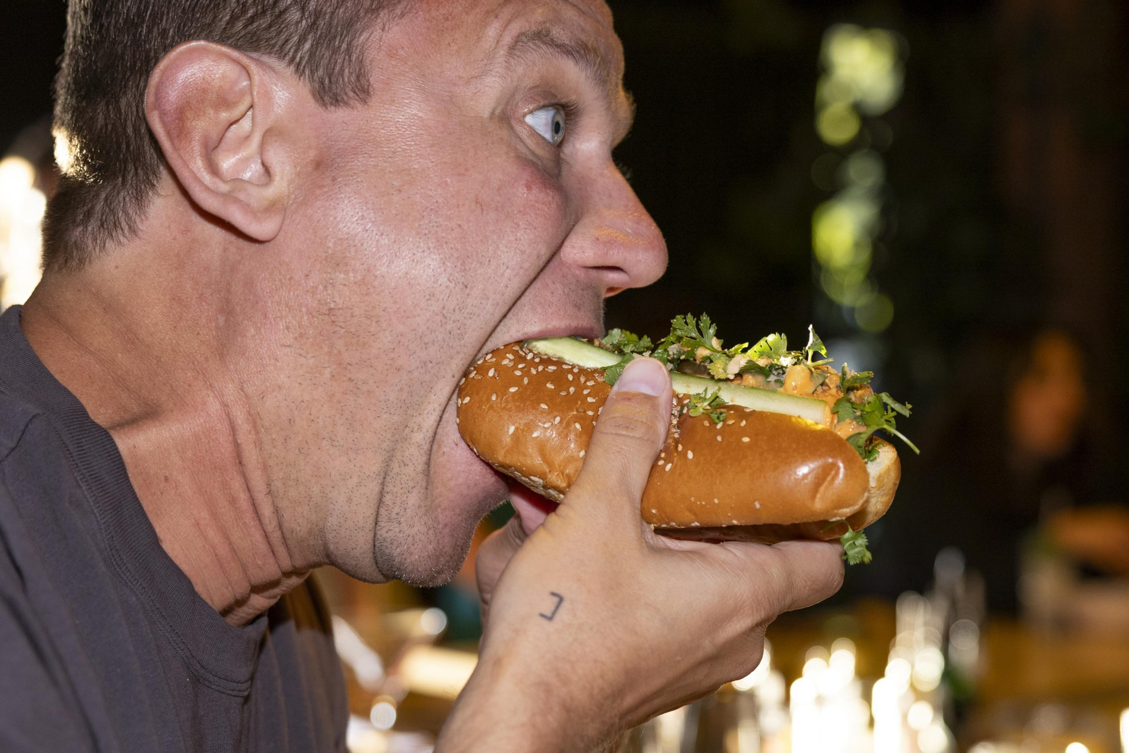 A man with a shocked expression is taking a large bite of a loaded sandwich on a sesame seed bun.