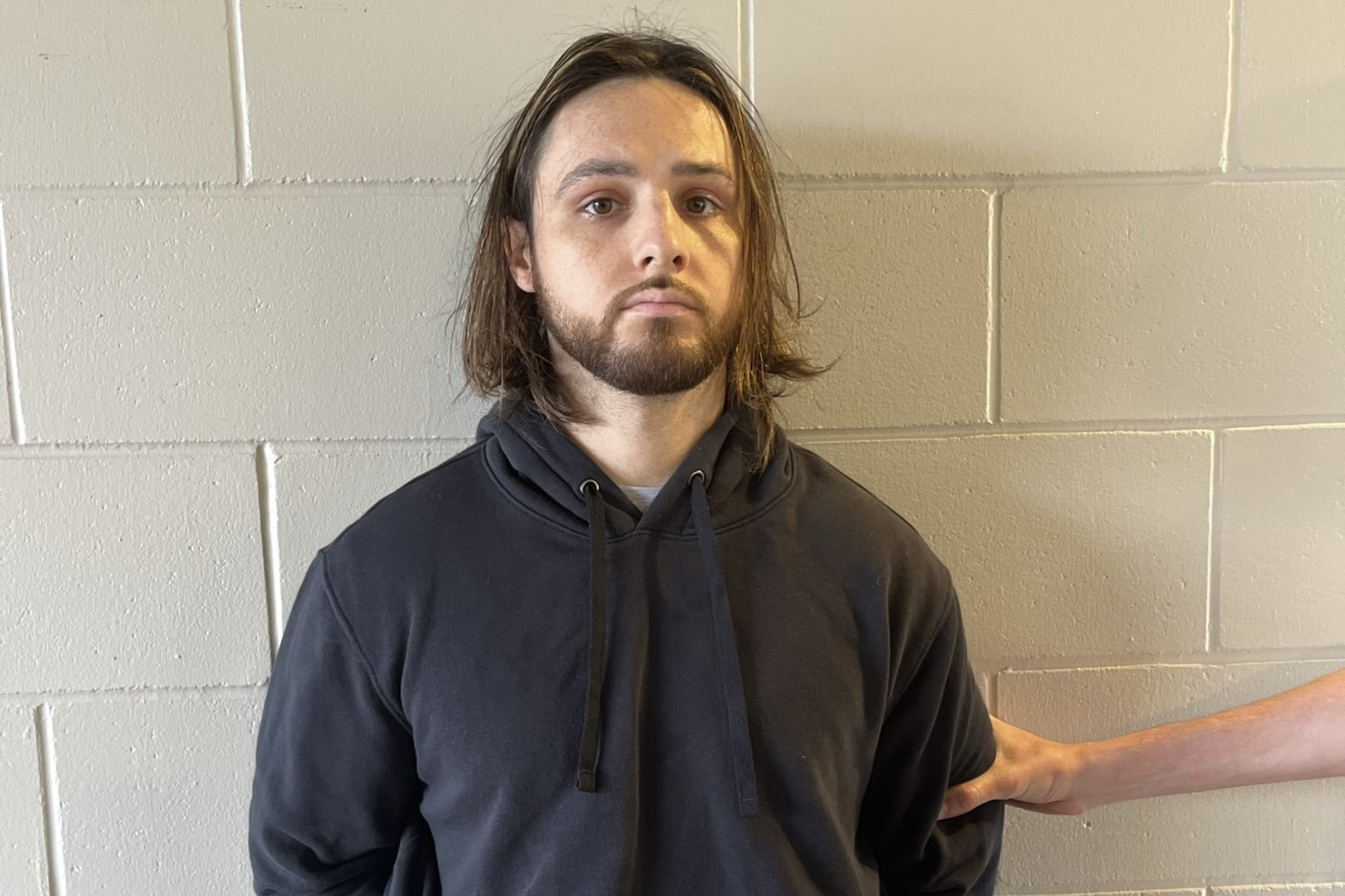 A man with shoulder-length hair and a beard wears a black hoodie and stands against a beige brick wall with his hands behind his back.