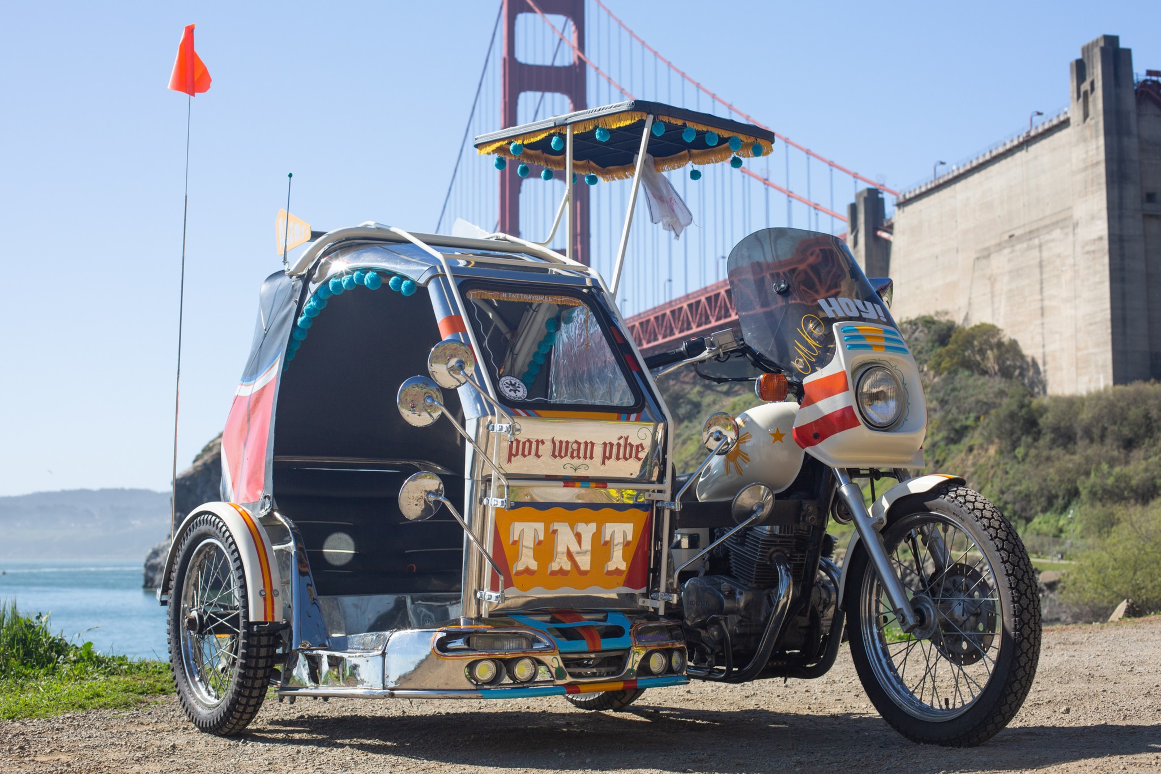 A colorful motorized tricycle with "TNT" on its side is parked by the water near the Golden Gate Bridge under a clear blue sky.