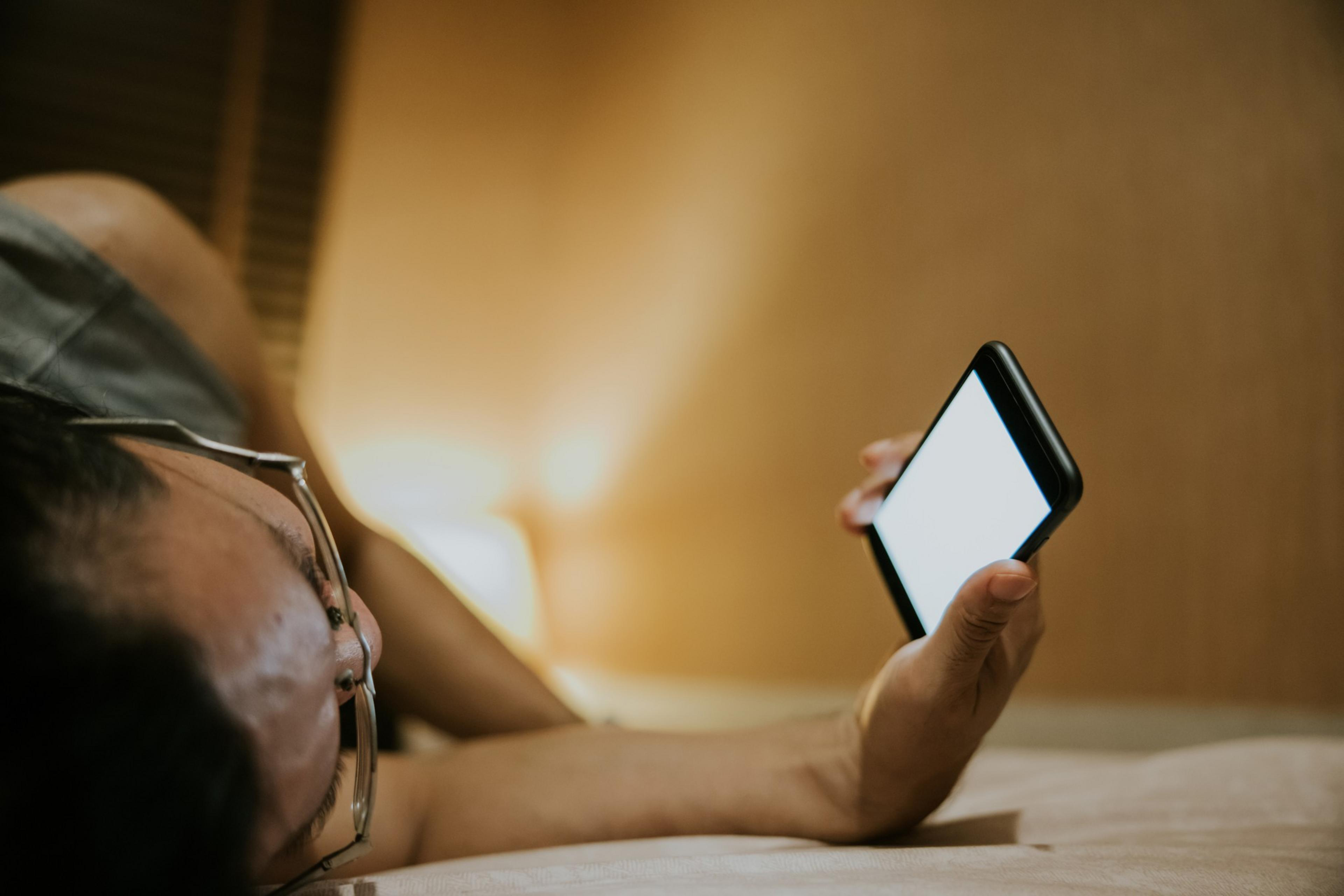 A person lying down is holding and looking at a smartphone with a blank white screen.