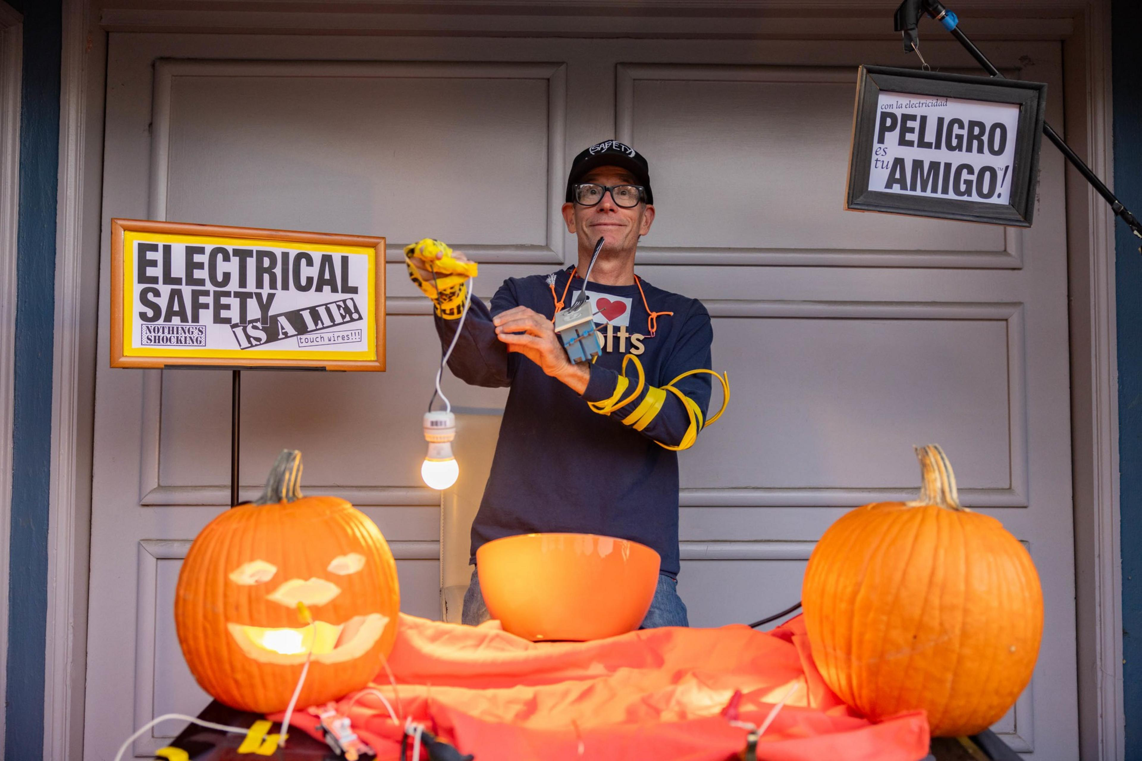 A person wearing gloves and a cap holds a dangling light bulb connected to electrical wires, with two pumpkins and warning signs about electrical safety nearby.