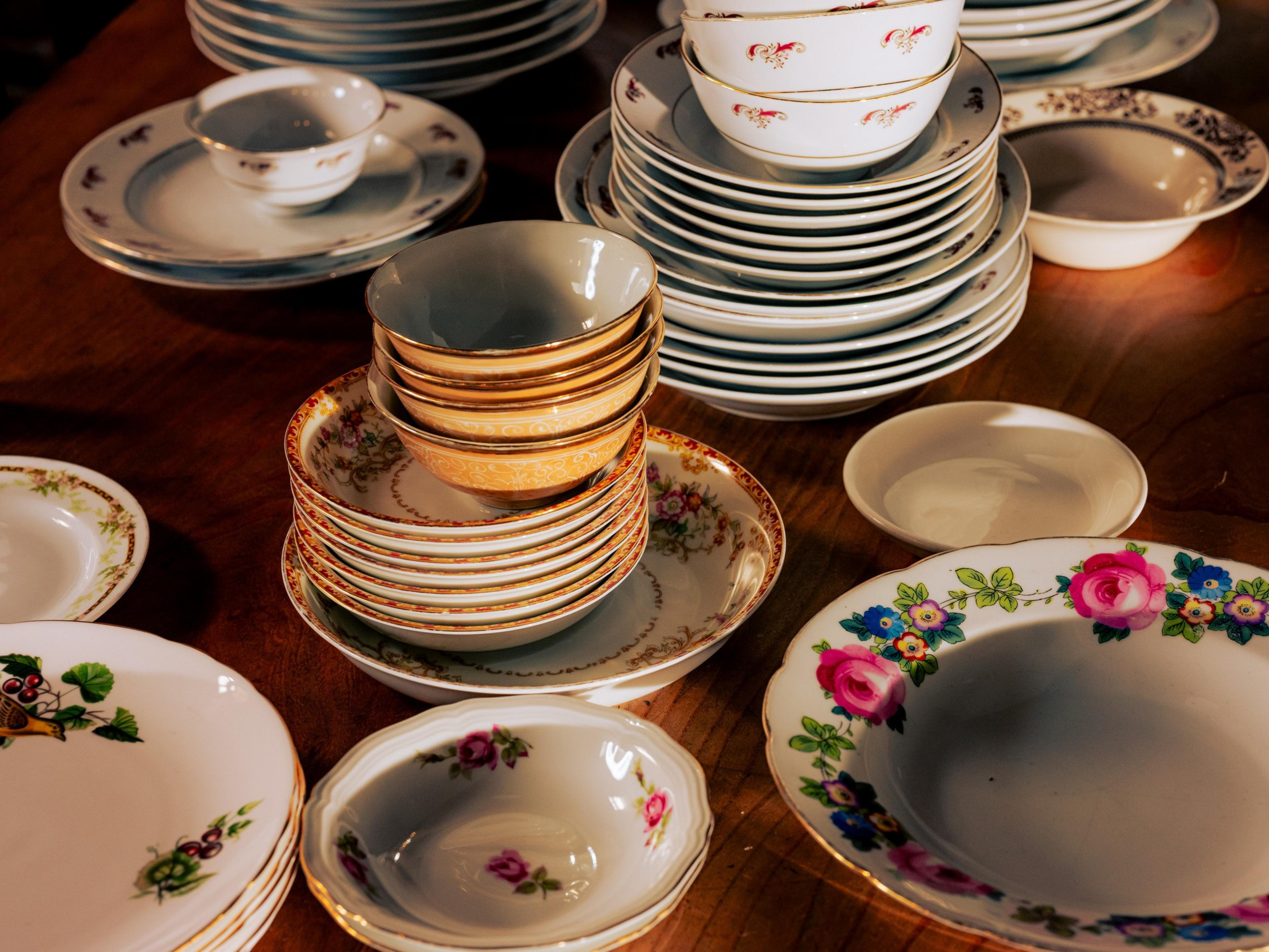 The image shows a wooden table covered with stacks of assorted patterned plates and bowls, featuring floral and decorative designs in various colors.