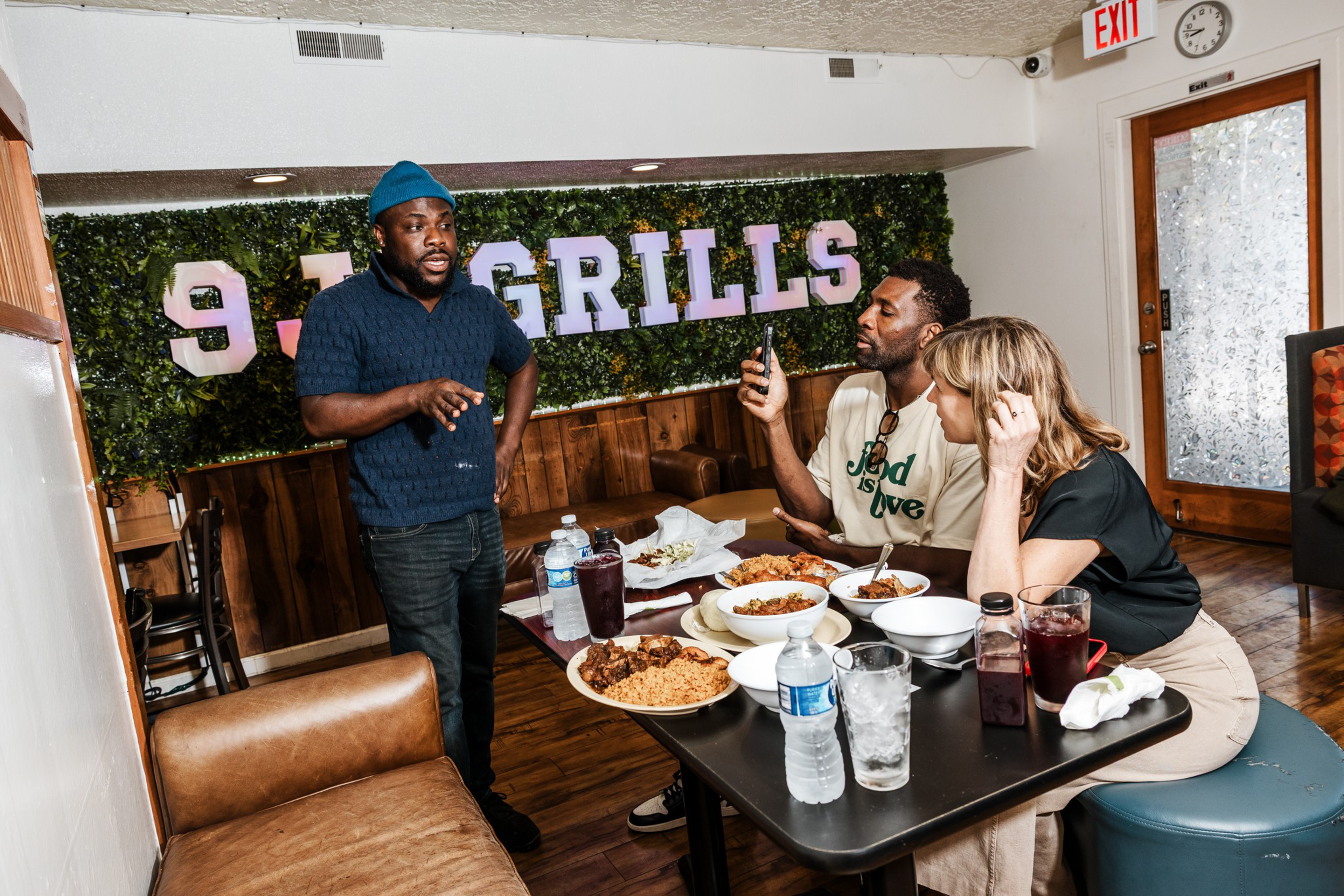 Two people sit eating a meal with rice and drinks at a table while a third person stands talking to them in a casual dining setting with “9.3 Grills” on the wall.