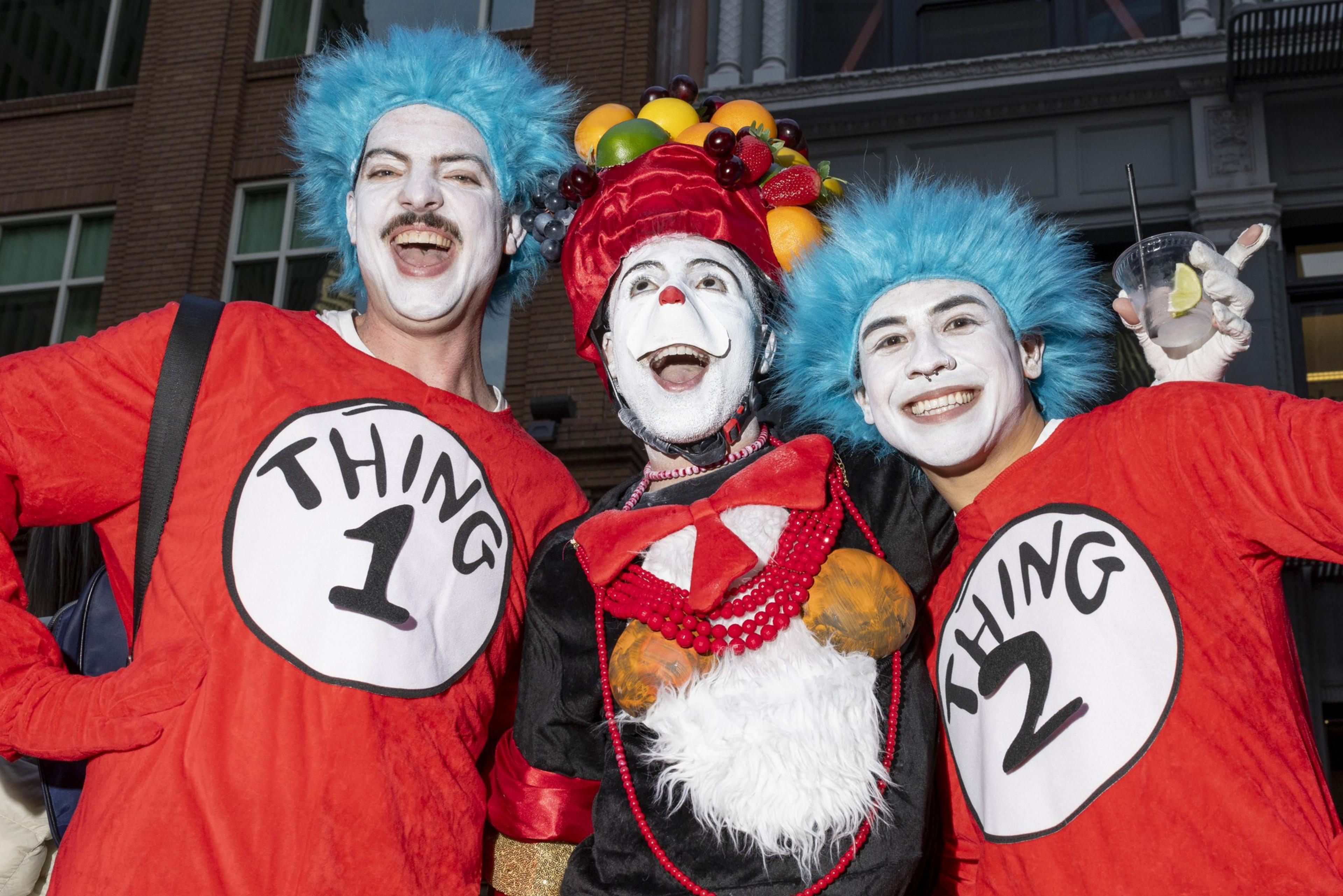 Three people in colorful costumes are smiling; two dressed as &quot;Thing 1&quot; and &quot;Thing 2&quot; with blue wigs, and one in a fruit-adorned hat and red outfit.