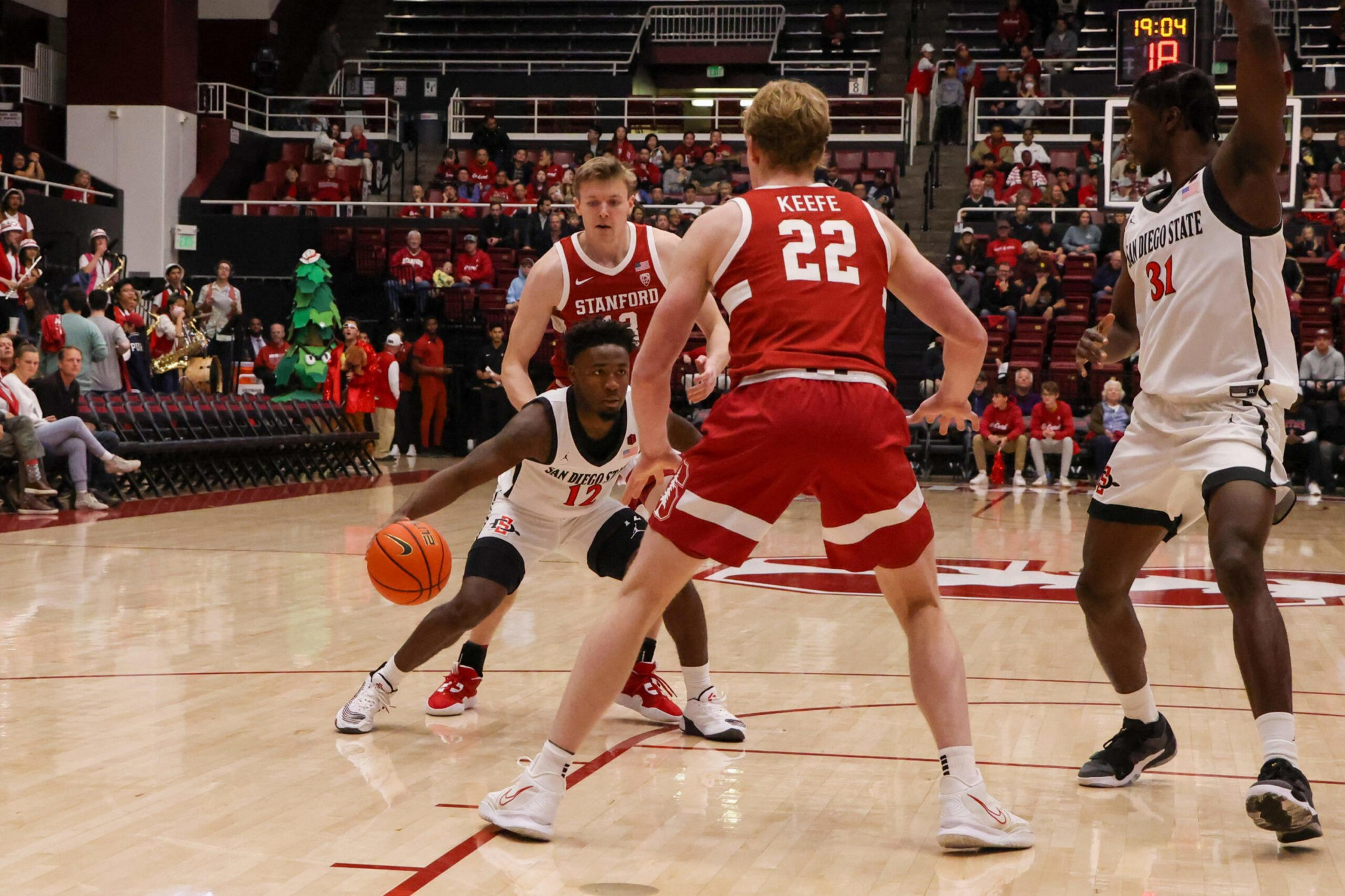 A San Diego State player dribbles the basketball while being guarded by a Stanford player wearing jersey number 22, with other players and spectators in the background.