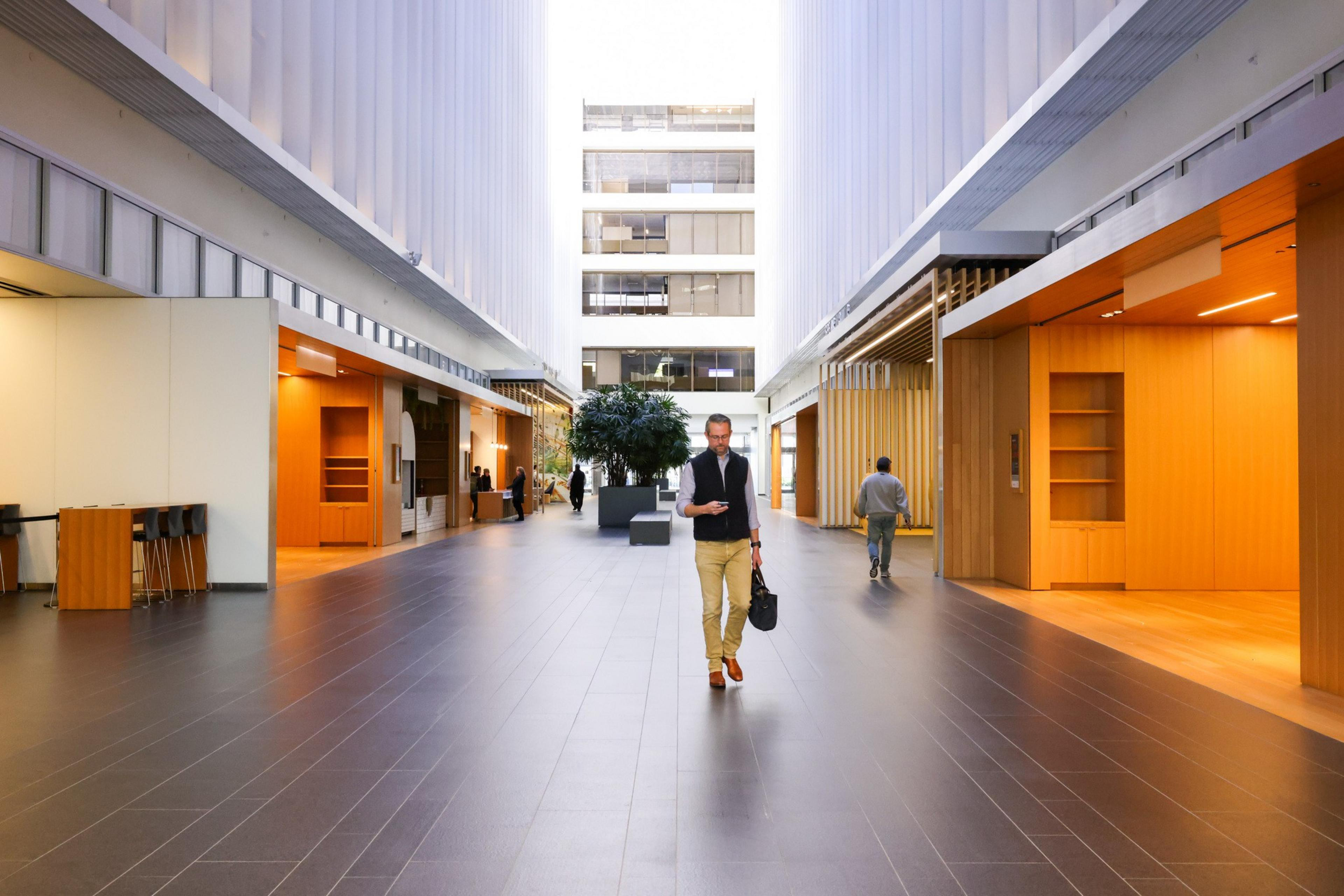 A man walks through a spacious, modern hallway with wood and white panel walls. Others are seen in the background near a large indoor plant.