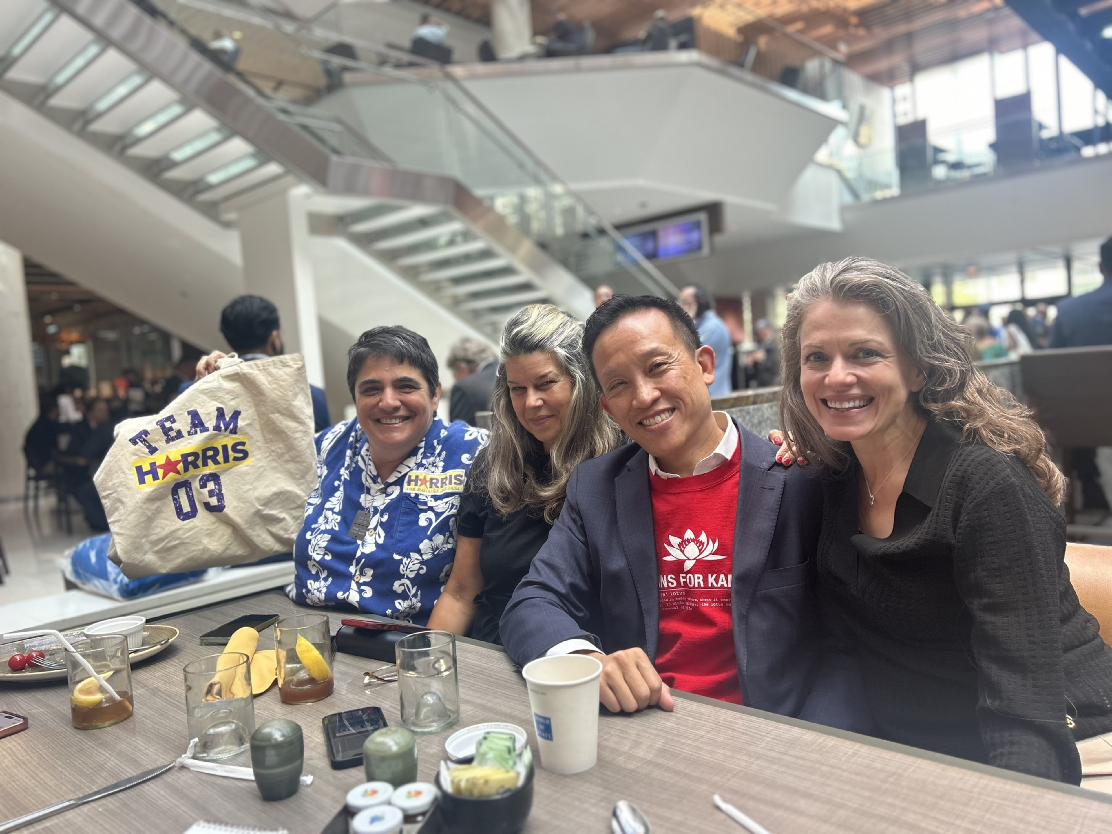 In a modern, indoor setting, four people sit at a table smiling, with one holding a "Team Harris 03" tote bag. Various drinks and snacks are on the table.