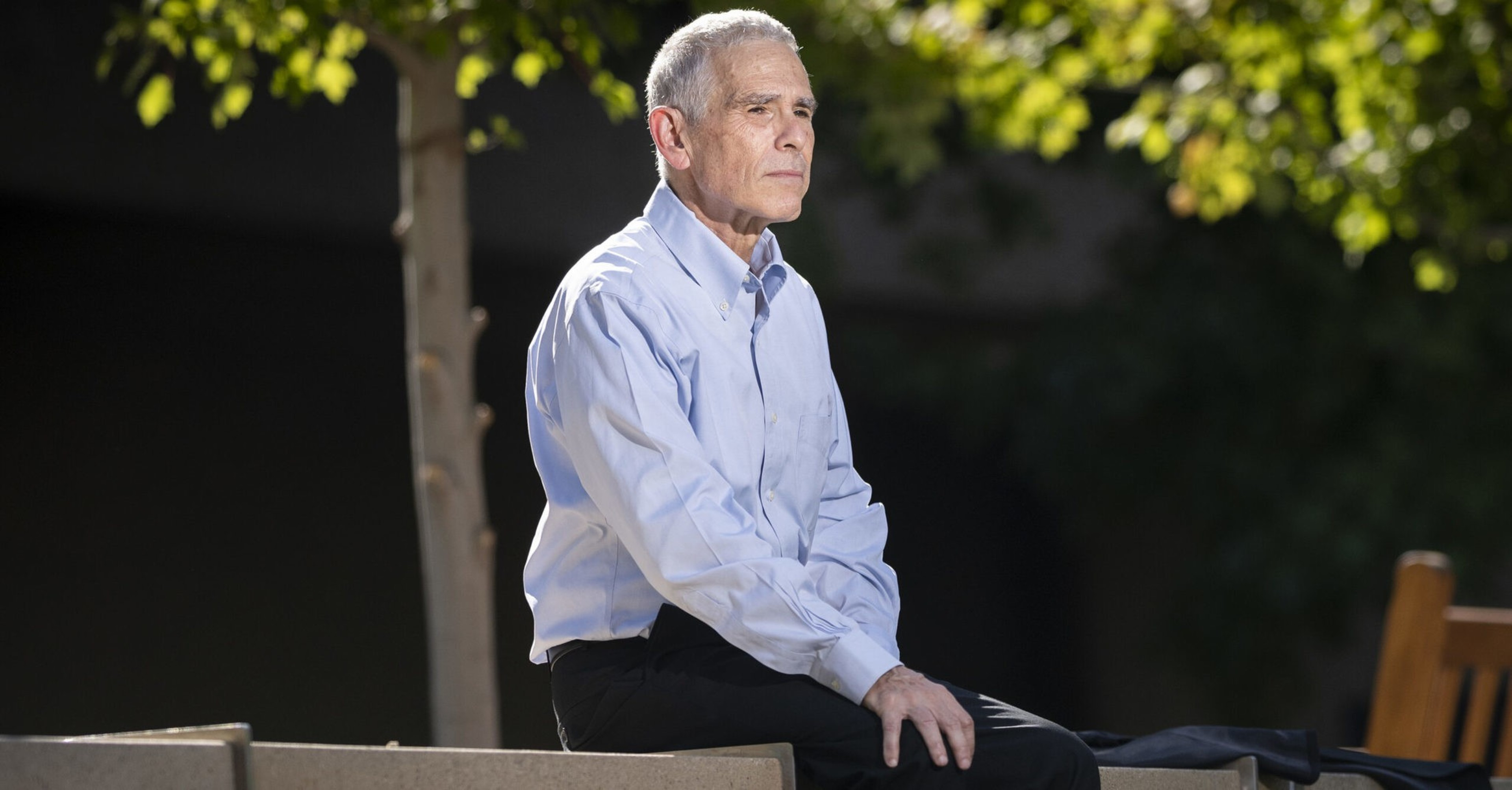 An elderly man with gray hair, wearing a light blue shirt and black pants, sits pensively outdoors on a concrete bench under a tree.