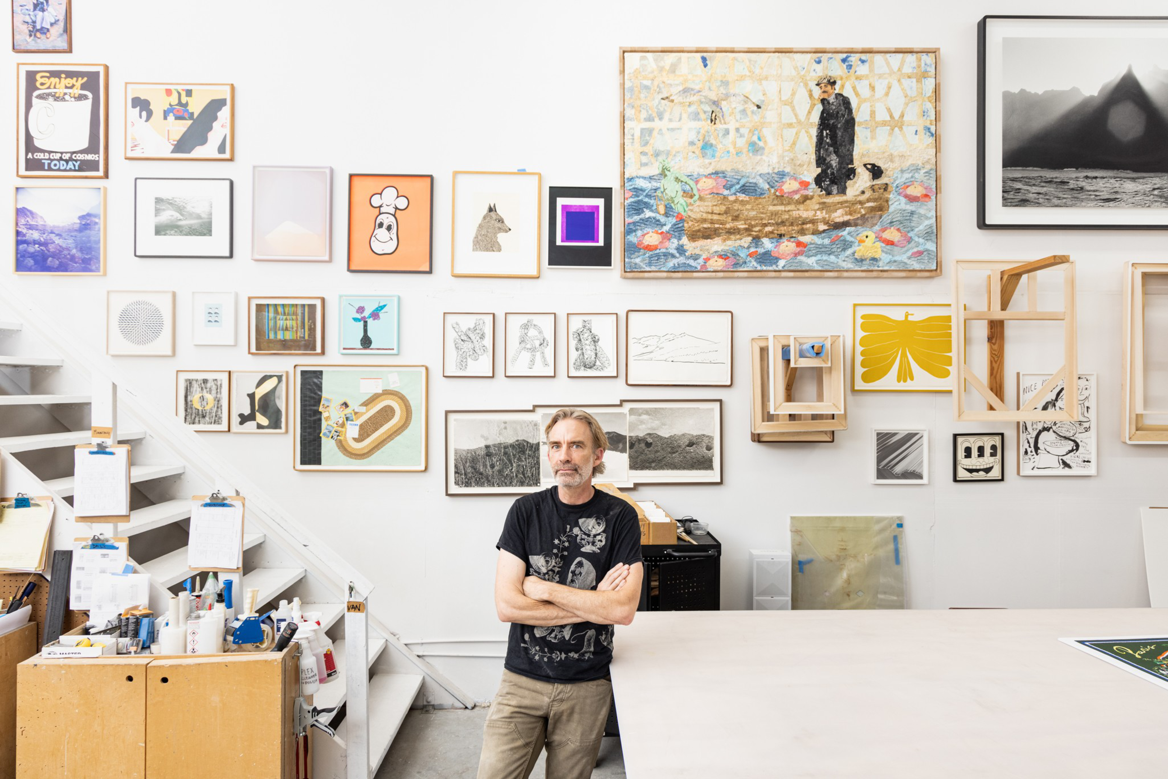 A man stands with folded arms in a studio filled with various framed artworks hung on the wall above a workspace and storage area.