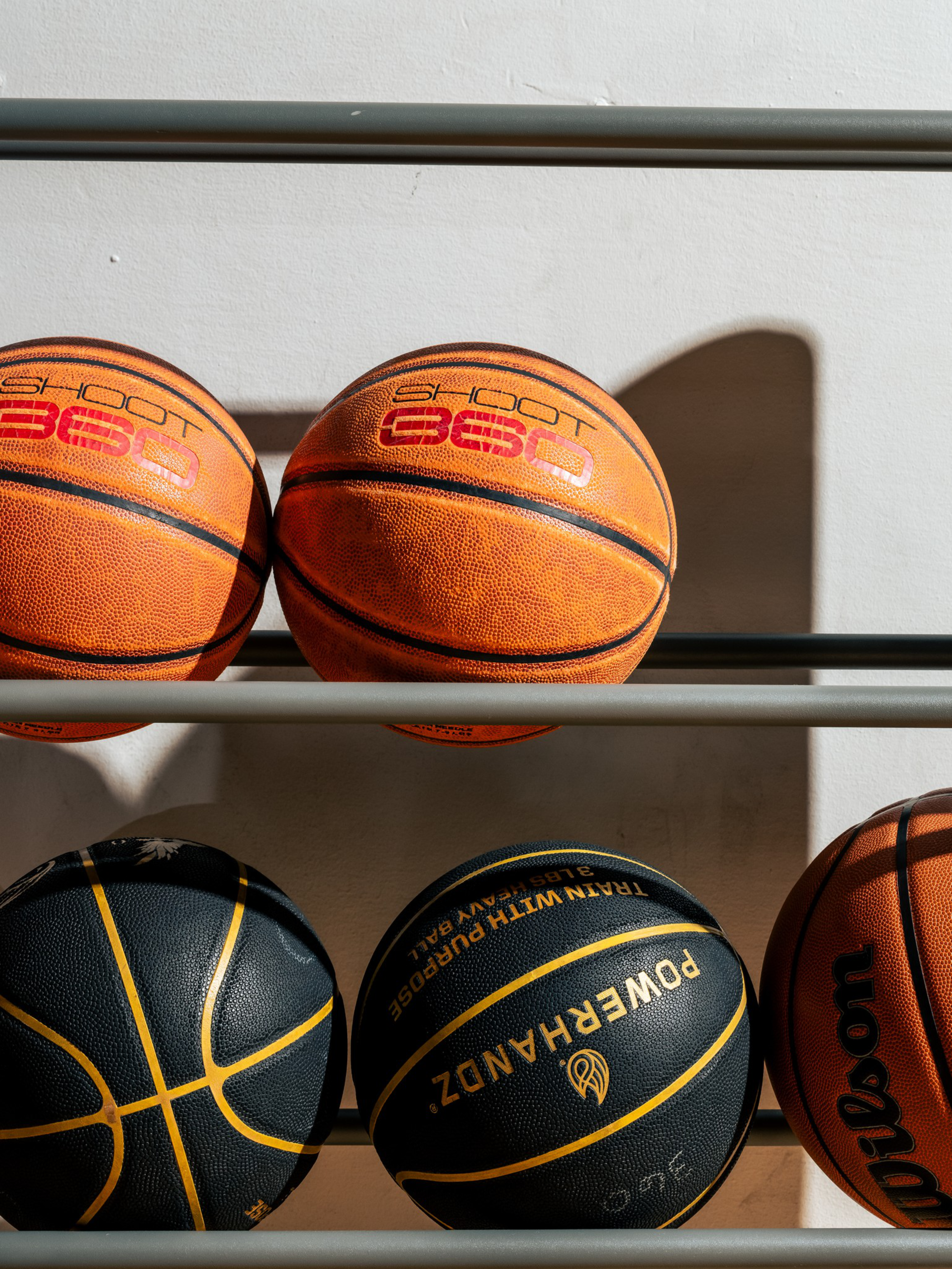 Two orange basketballs with “SHOOT 360" on the top row and three basketballs, including two black and one orange Wilson, on the bottom row of a rack.