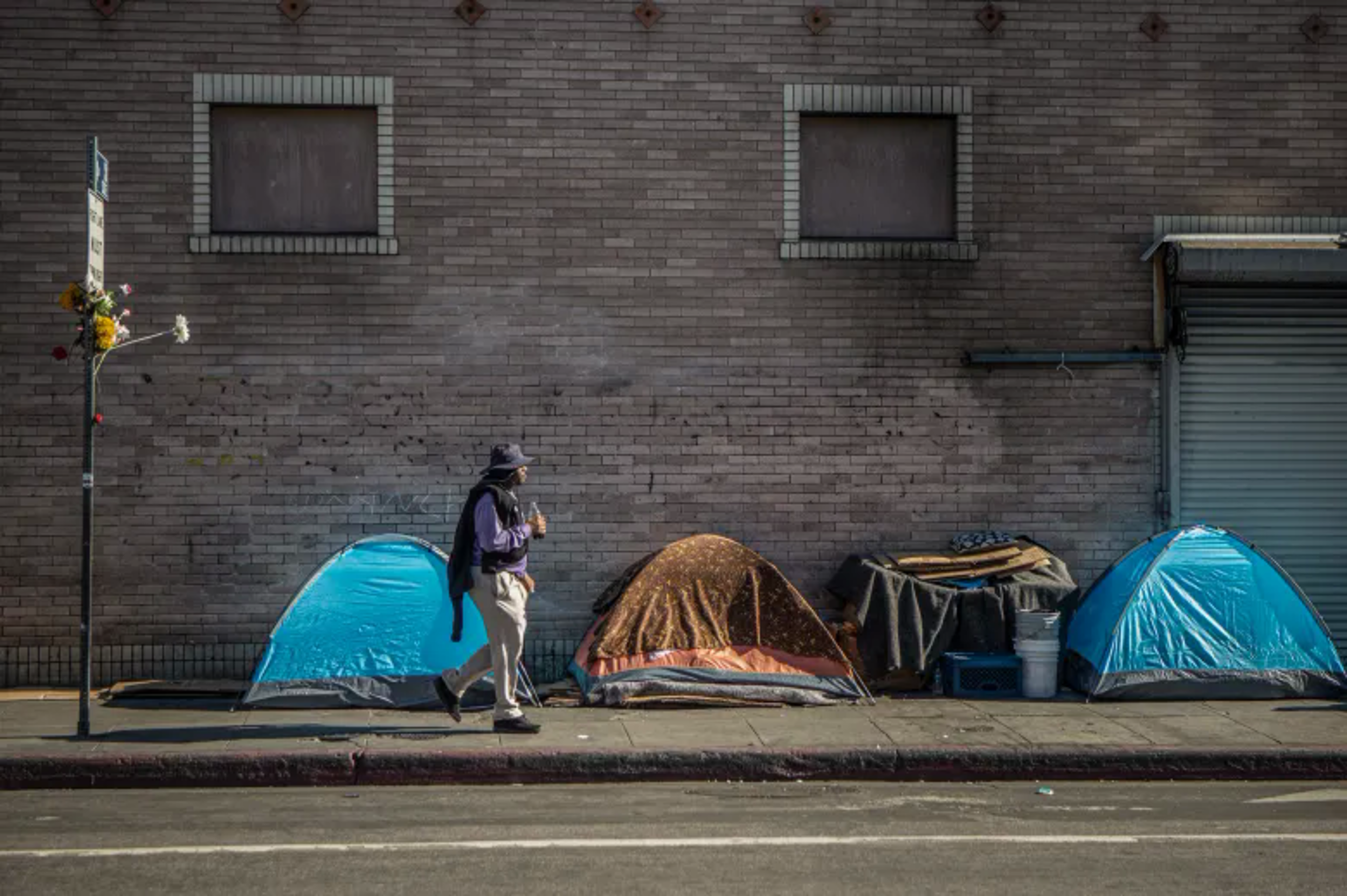 Three small tents and a pile of belongings line a sidewalk against a brick wall, with a person walking by wearing a hat and scarf.