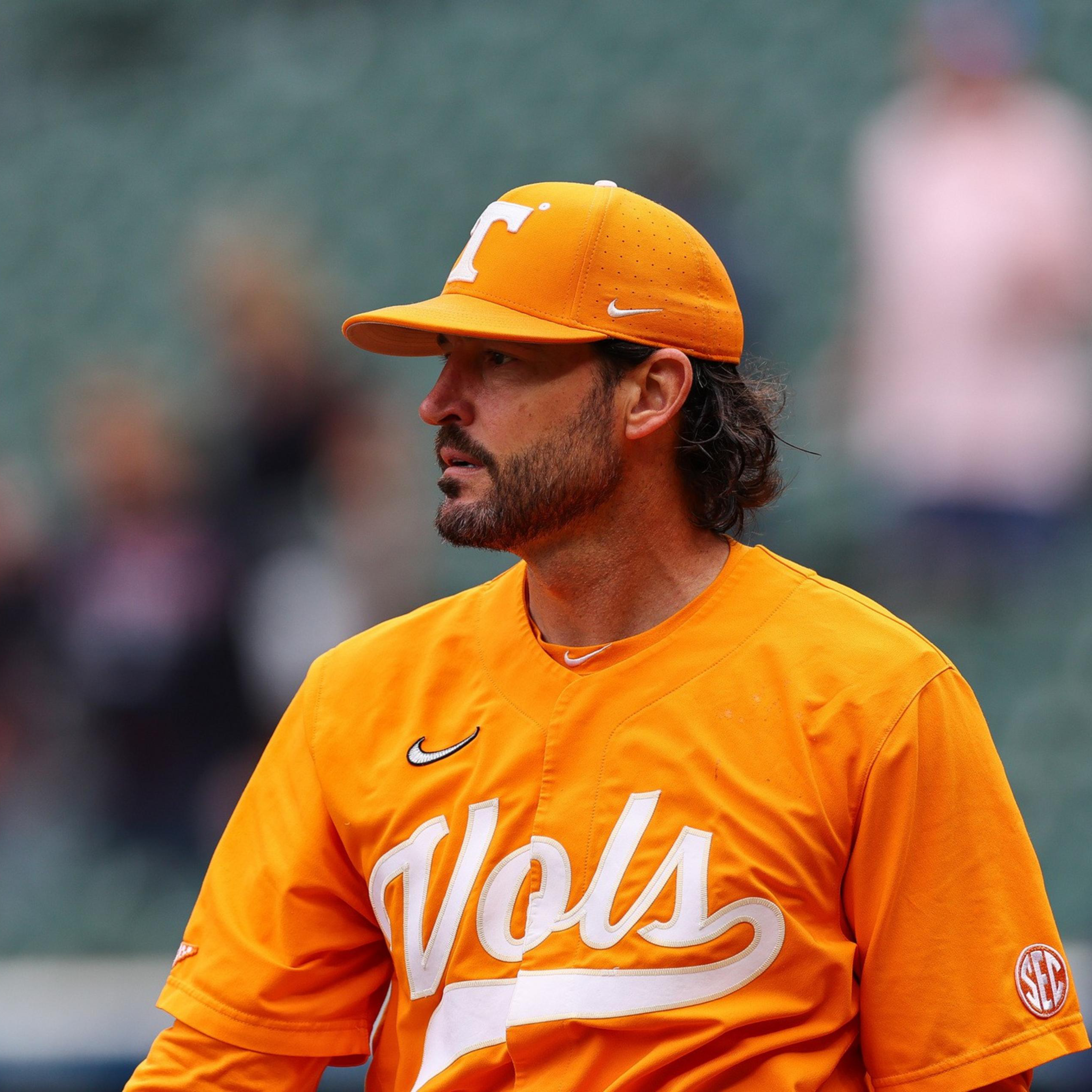 A man wearing an orange baseball uniform and cap with "Vols" written on the jersey stands looking to the side in a stadium with blurred spectators in the background.