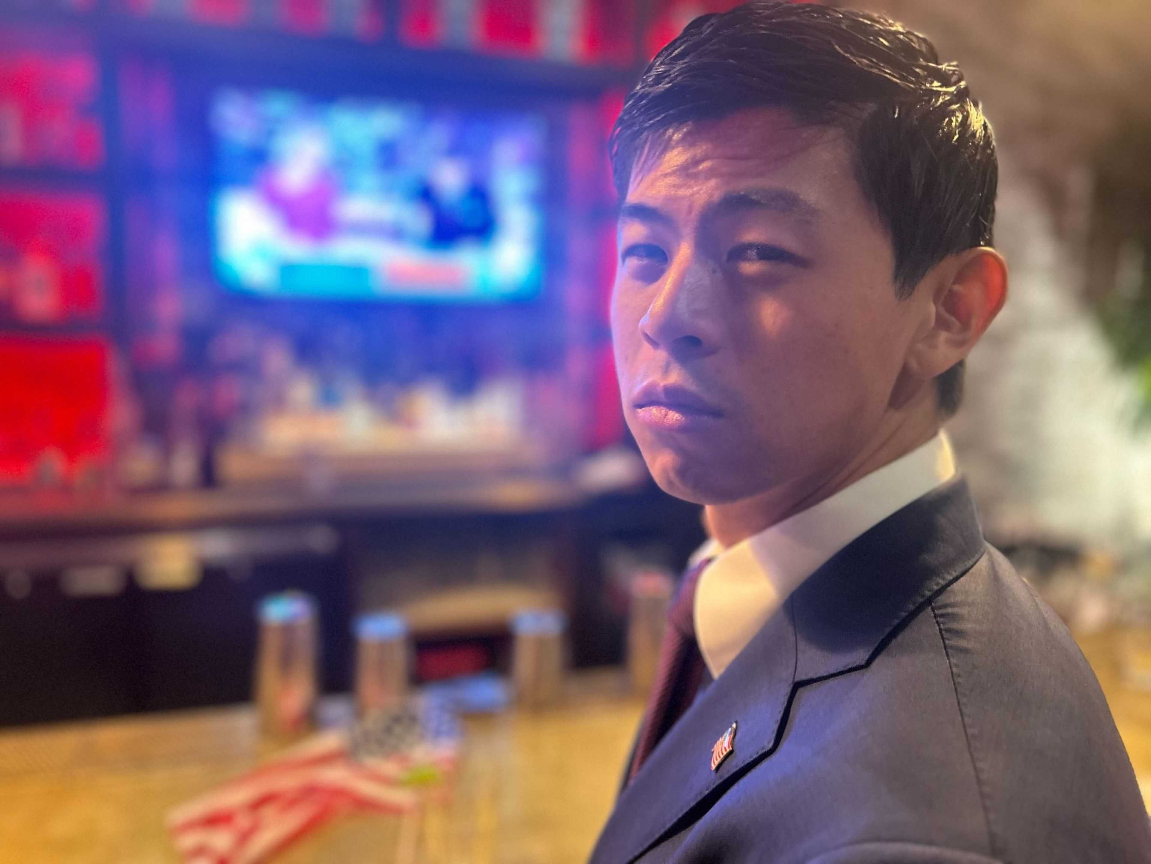 A man in a suit with an American flag pin looks intently at the camera. Behind him is a blurred bar with a bright TV screen showing figures.
