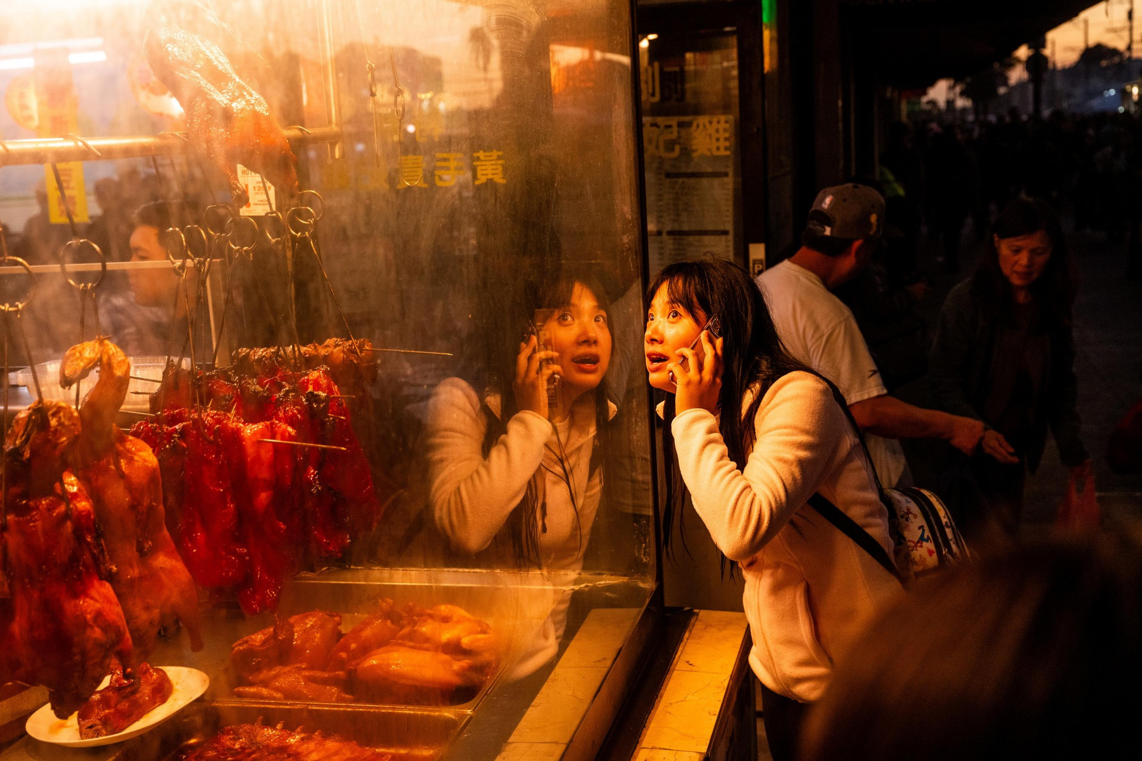 A woman talks on the phone while peering into a window displaying roasted meats. Her reflection is visible, and people walk by on the dimly lit street.